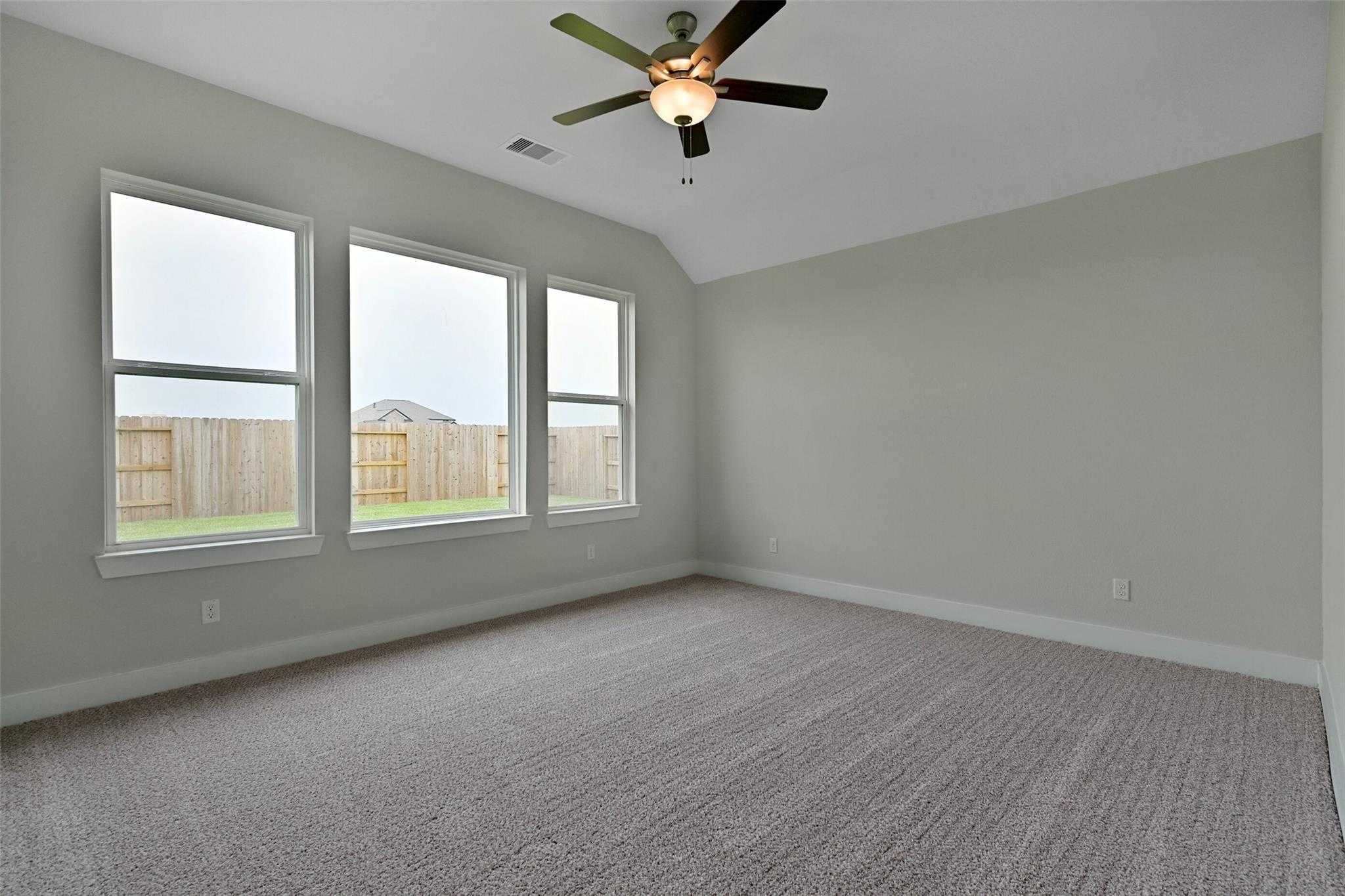 Bright bedroom with light gray walls, three large windows, ceiling fan, and backyard view in Davidson Homes The Edward A, Lago Mar, Texas City