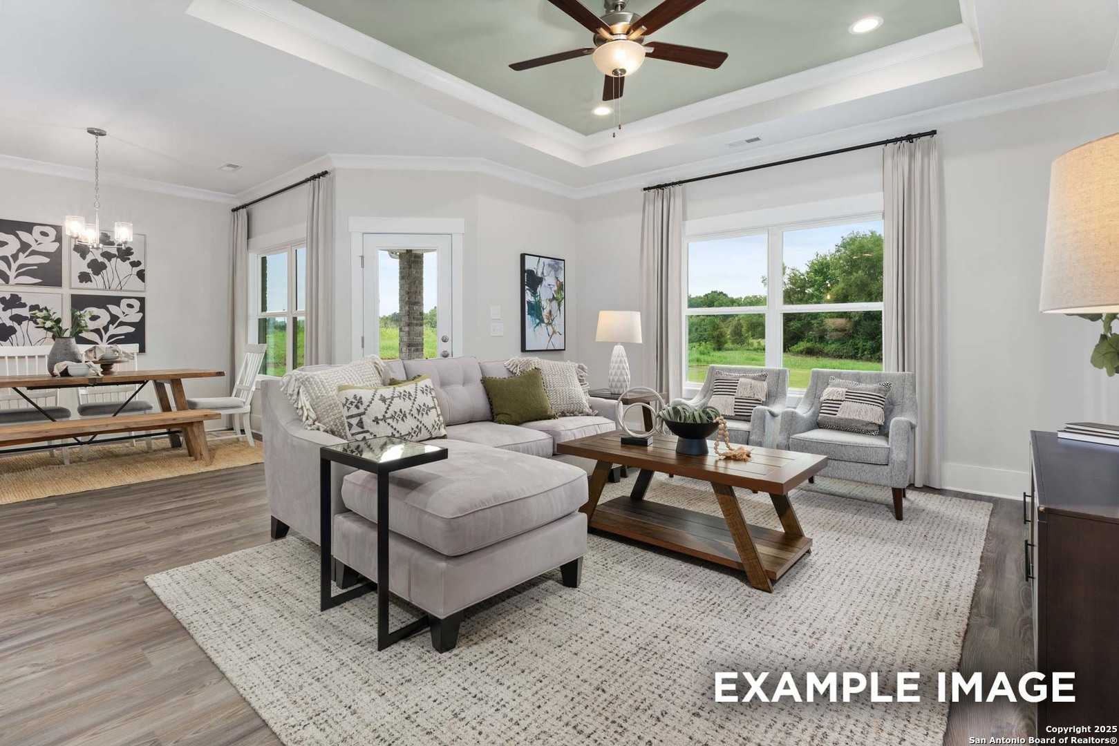 Bright open-concept living room with gray sectional sofa, wooden coffee table, dining bench, and large windows overlooking trees in Davidson Homes The Daphne H, Seguin, Texas