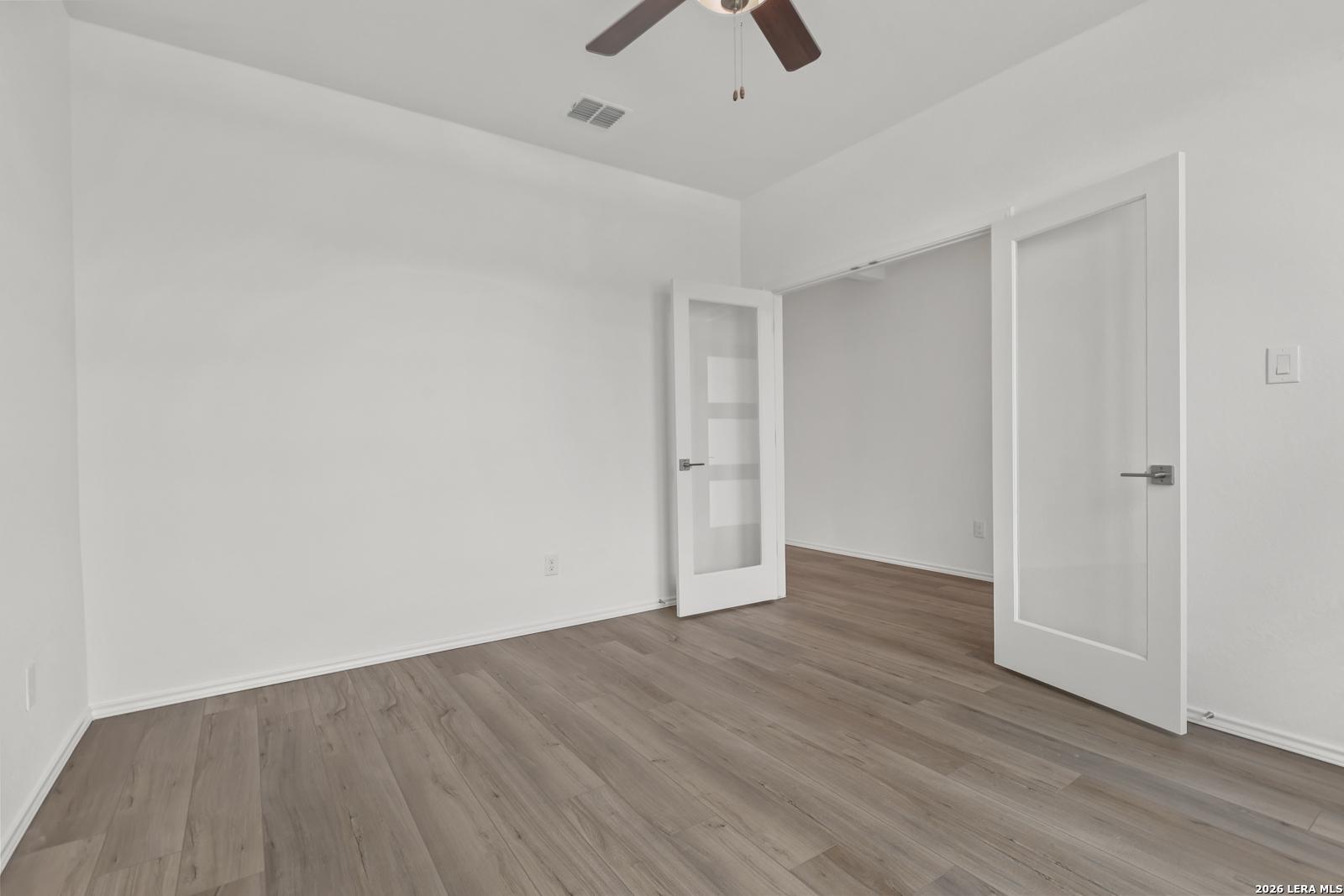 Bright empty bedroom with white walls, gray luxury vinyl plank floors, ceiling fan, and frosted glass door in The Glenwood E, Elmendorf, Texas