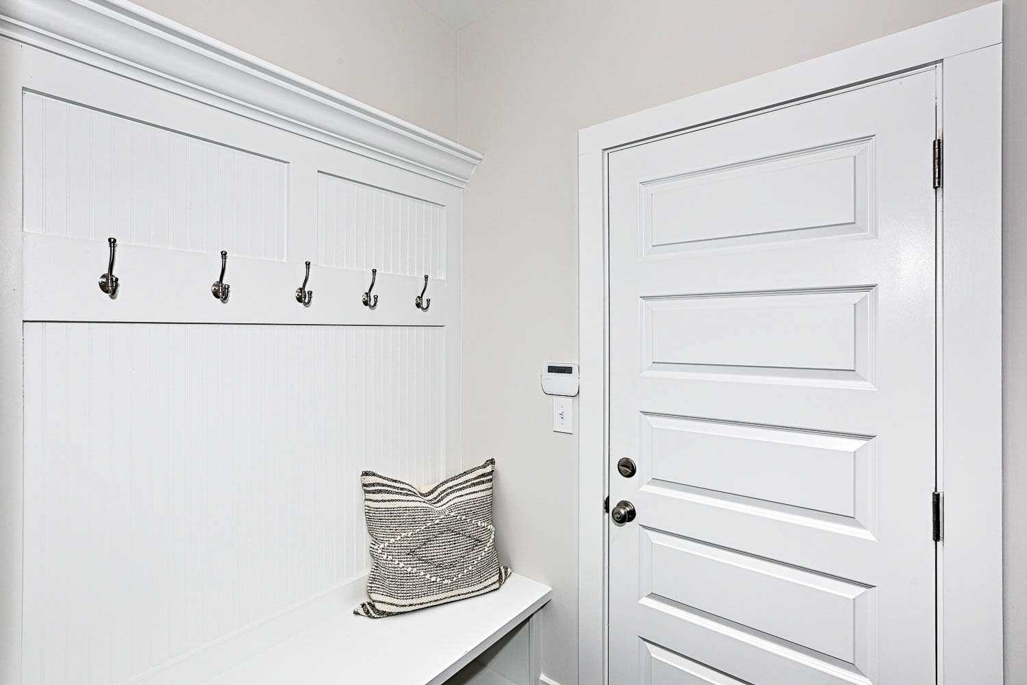 Spacious mudroom in The Finleigh home with white cabinetry, bronze coat hooks, built-in bench, patterned pillow, and paneled door