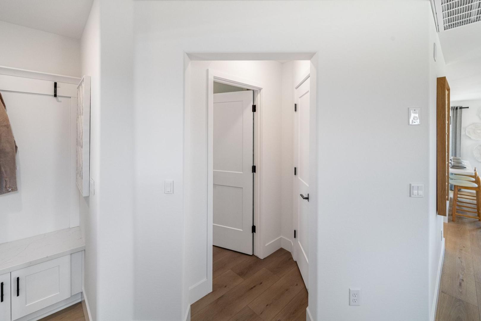Spacious mudroom in The Wilmington showcasing white walls, coat hooks with tan jacket, built-in bench, and open doorway to wooden-floored dining area