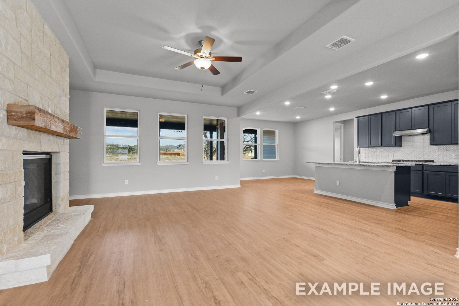 Open-concept living area in The Oxford H featuring a stone fireplace, tray ceiling, and modern kitchen with dark cabinetry.