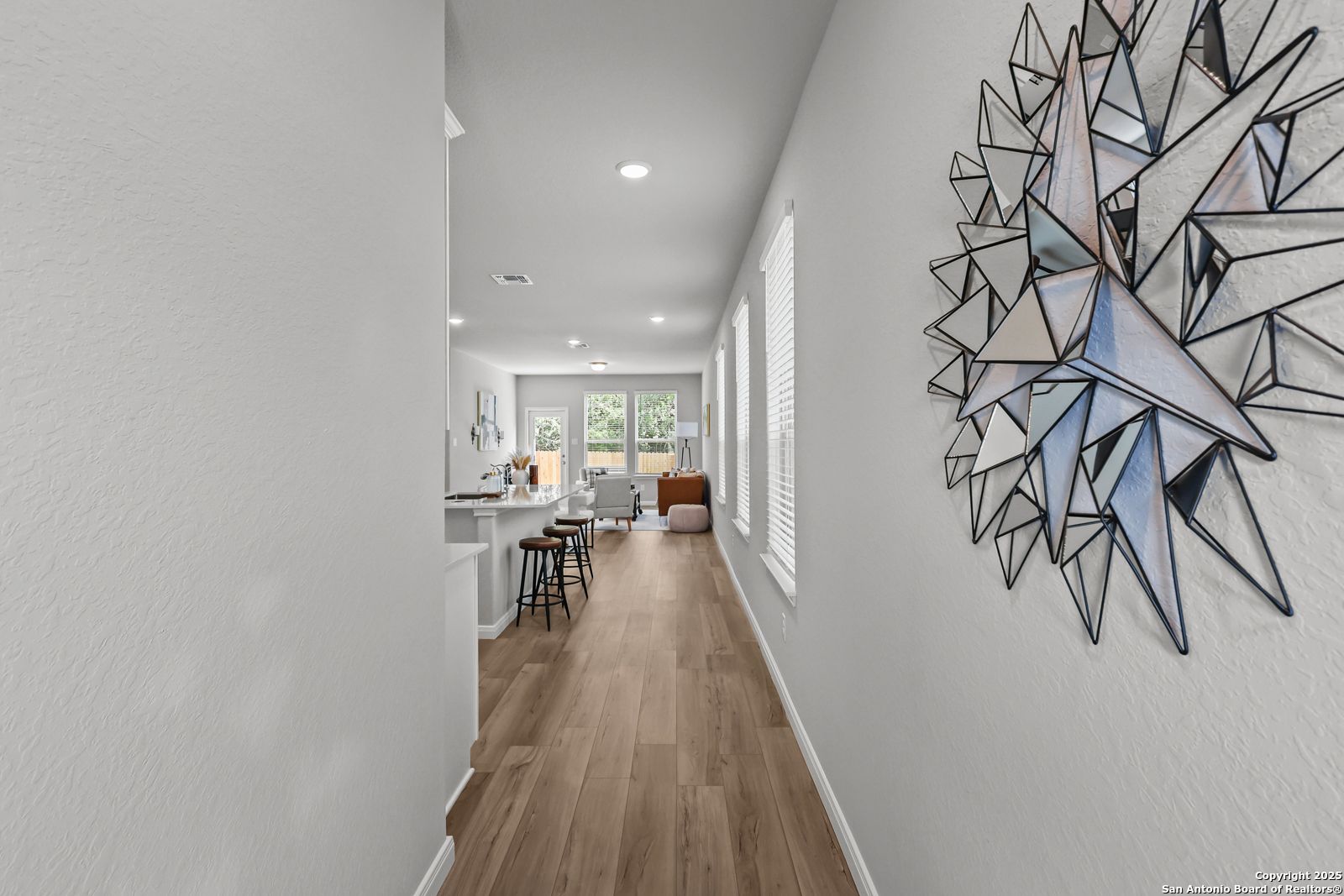 Long hallway with geometric wall art opening to modern white kitchen and breakfast bar in Davidson Homes The Gillian C, San Antonio