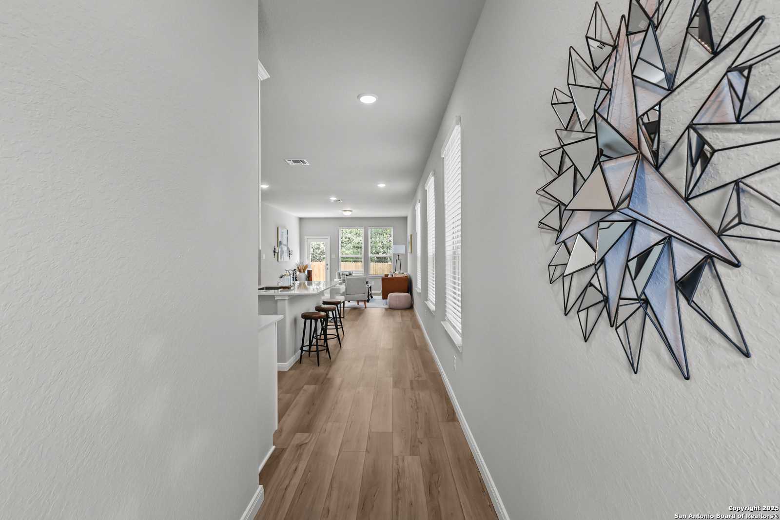 Long hallway with geometric wall art opening to modern white kitchen and breakfast bar in Davidson Homes The Gillian C, San Antonio