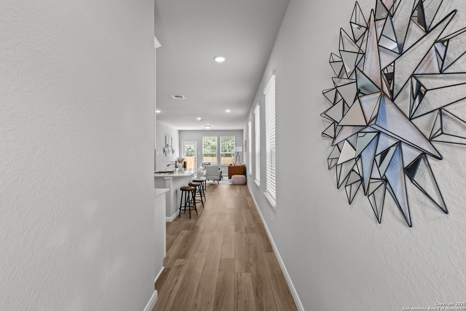 Long hallway with geometric wall art opening to modern white kitchen and breakfast bar in Davidson Homes The Gillian C, San Antonio