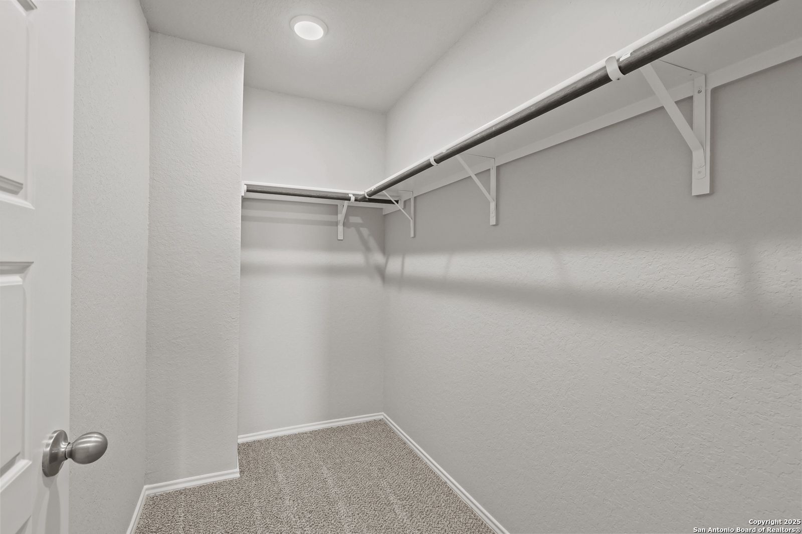 Spacious walk-in closet with built-in white shelves and hanging rods on light walls, gray carpet floor in Davidson Homes The Trinity D, San Antonio