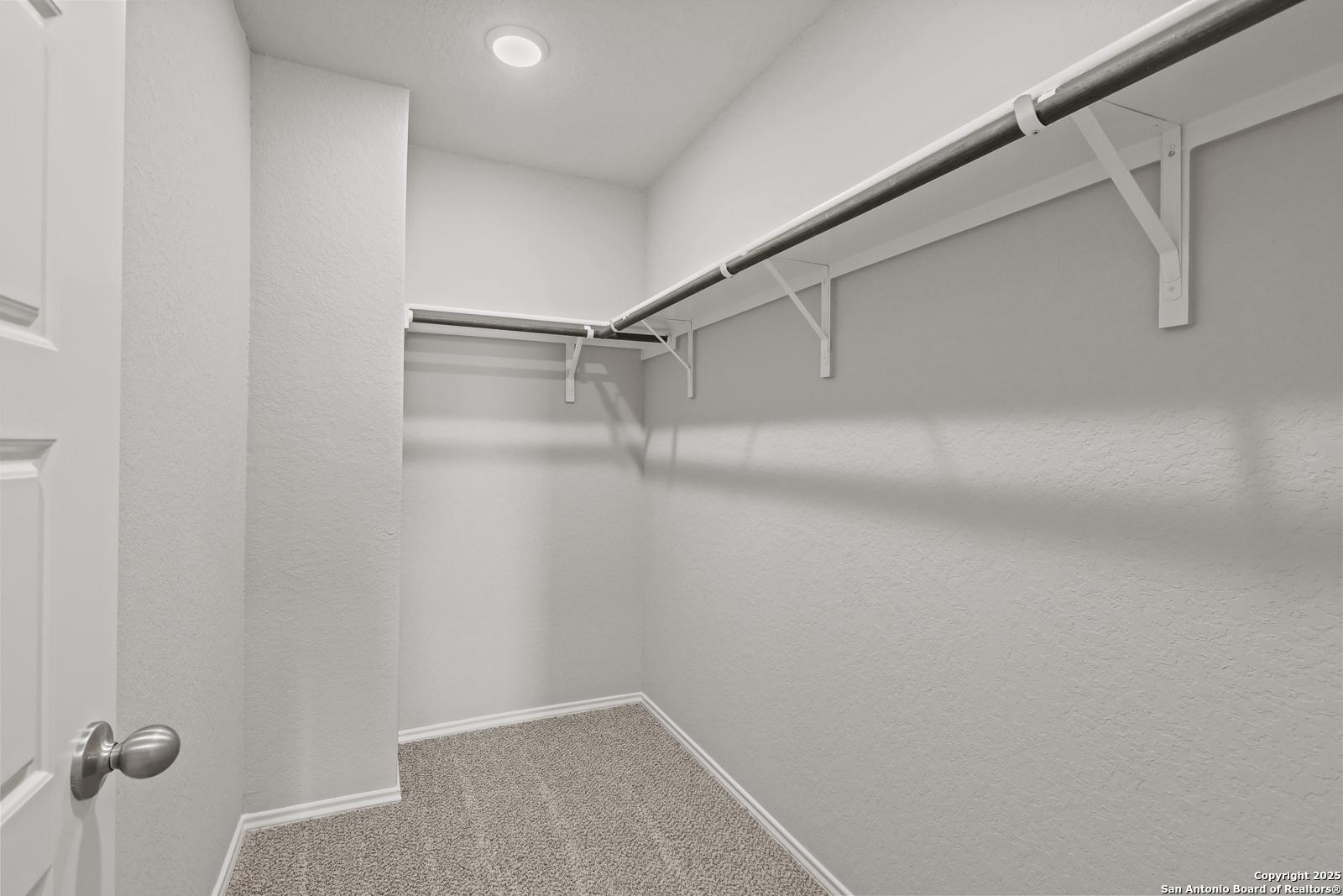 Spacious walk-in closet with built-in white shelves and hanging rods on light walls, gray carpet floor in Davidson Homes The Trinity D, San Antonio