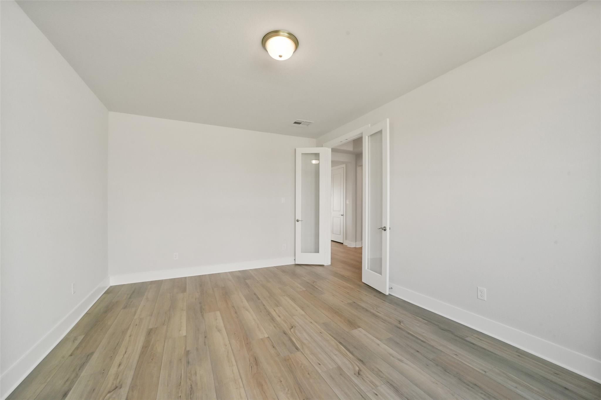 Bright bedroom with light hardwood floors, white walls, and open French doors in Davidson Homes The Zion A, Lago Mar, Texas City