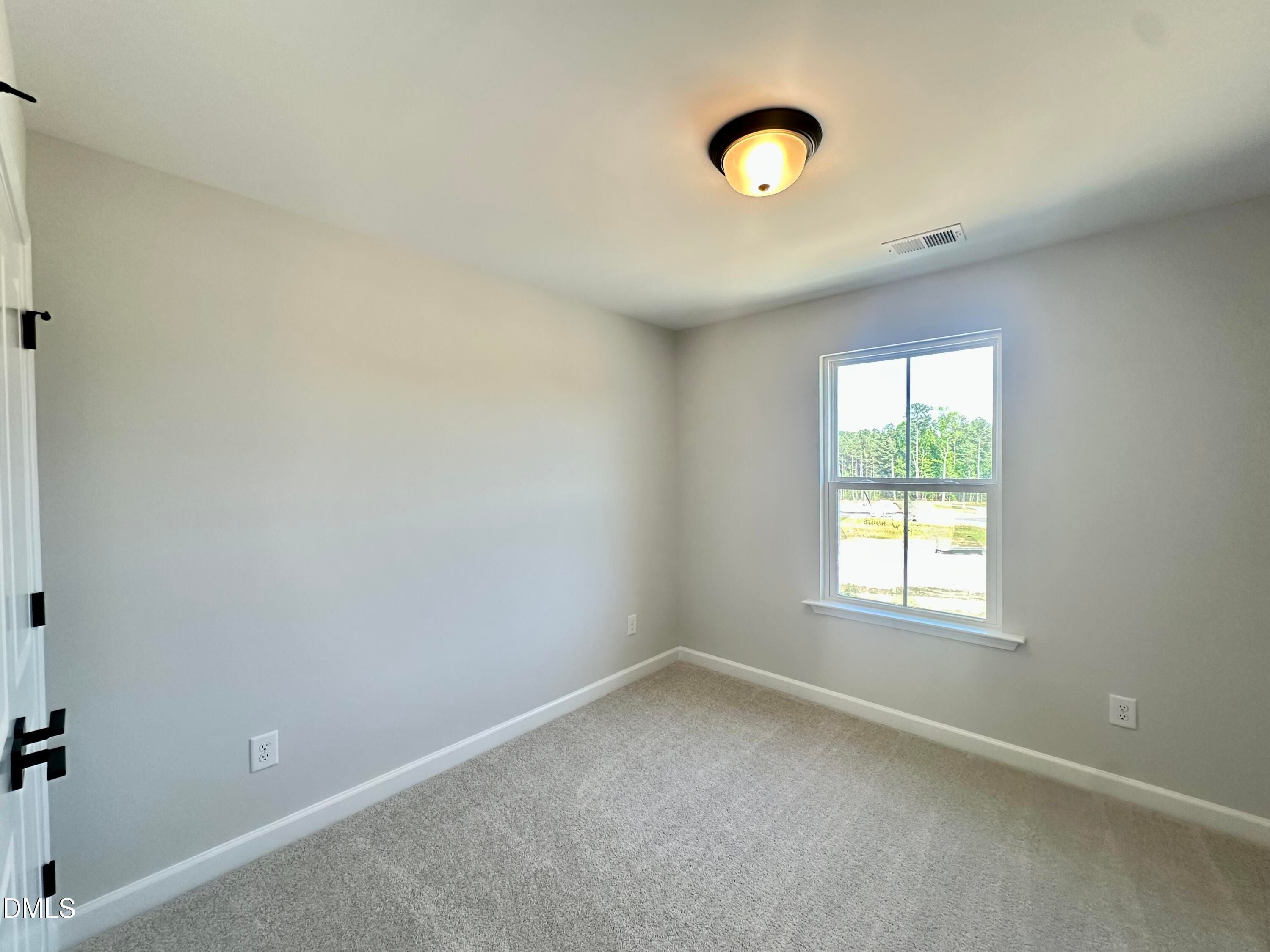 Bright empty bedroom with gray walls, neutral carpet, ceiling light, and wooded window view in Davidson Homes The Avery, Knightdale NC