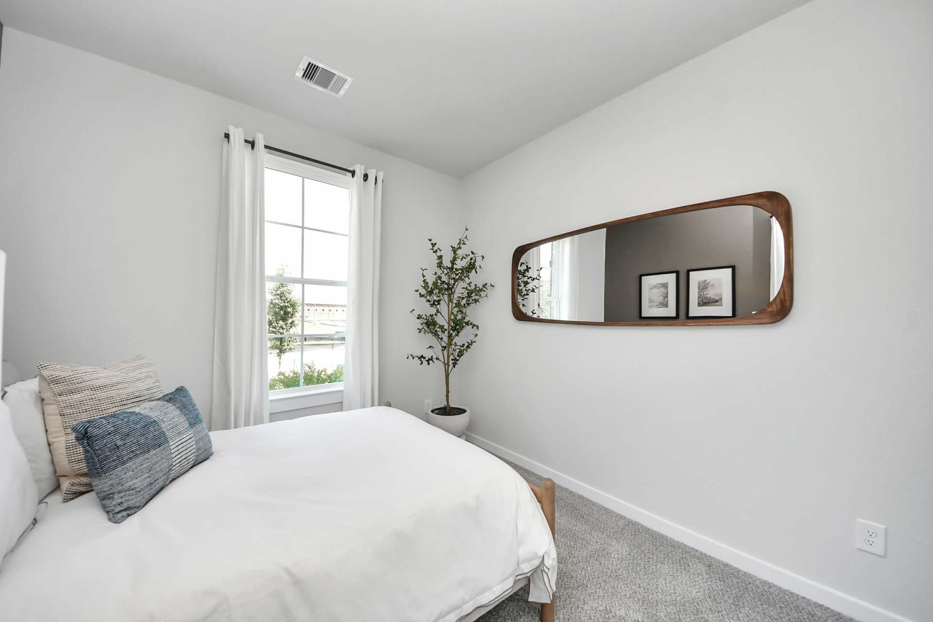 Bright bedroom with modern decor at Enclave at Newport in Crosby, TX, featuring a cozy bed, potted plant, and wall mirror.