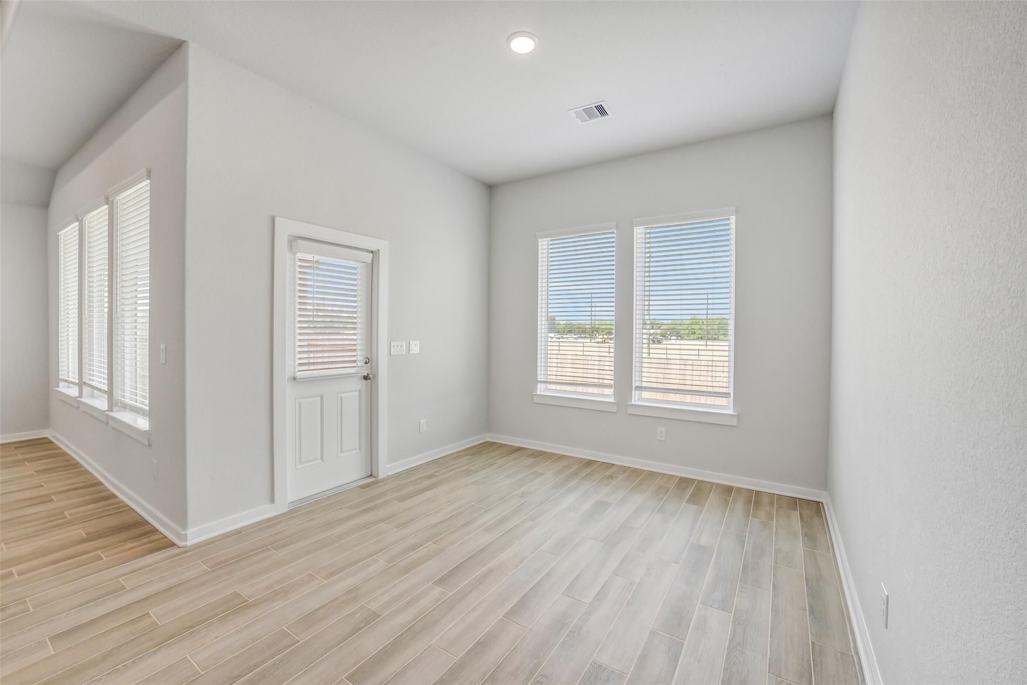 Bright corner room with gray walls, large blinds-covered windows, white door, and light wood flooring in Davidson Homes Acadia C, Magnolia TX