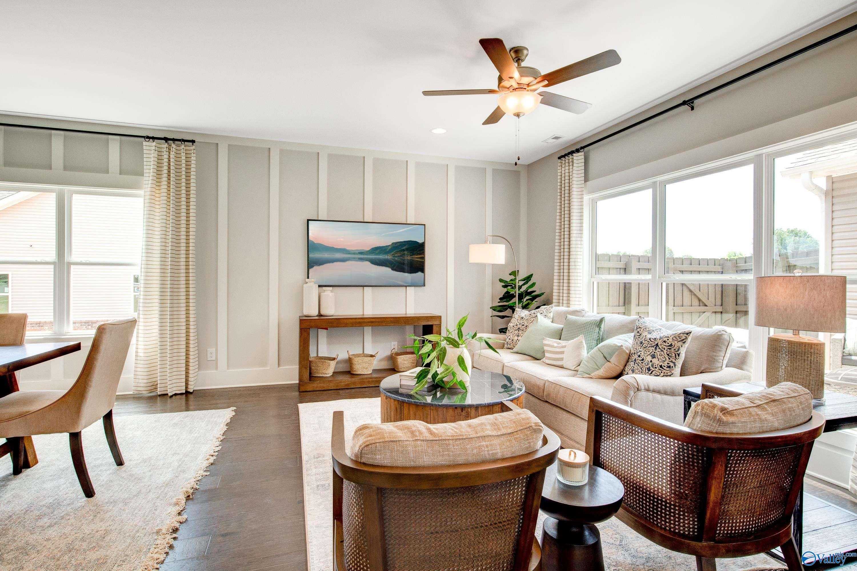 Cozy living room with beige walls, large windows, ceiling fan, plush sofa, armchairs, and plants in Davidson Homes Covington C, Decatur, AL
