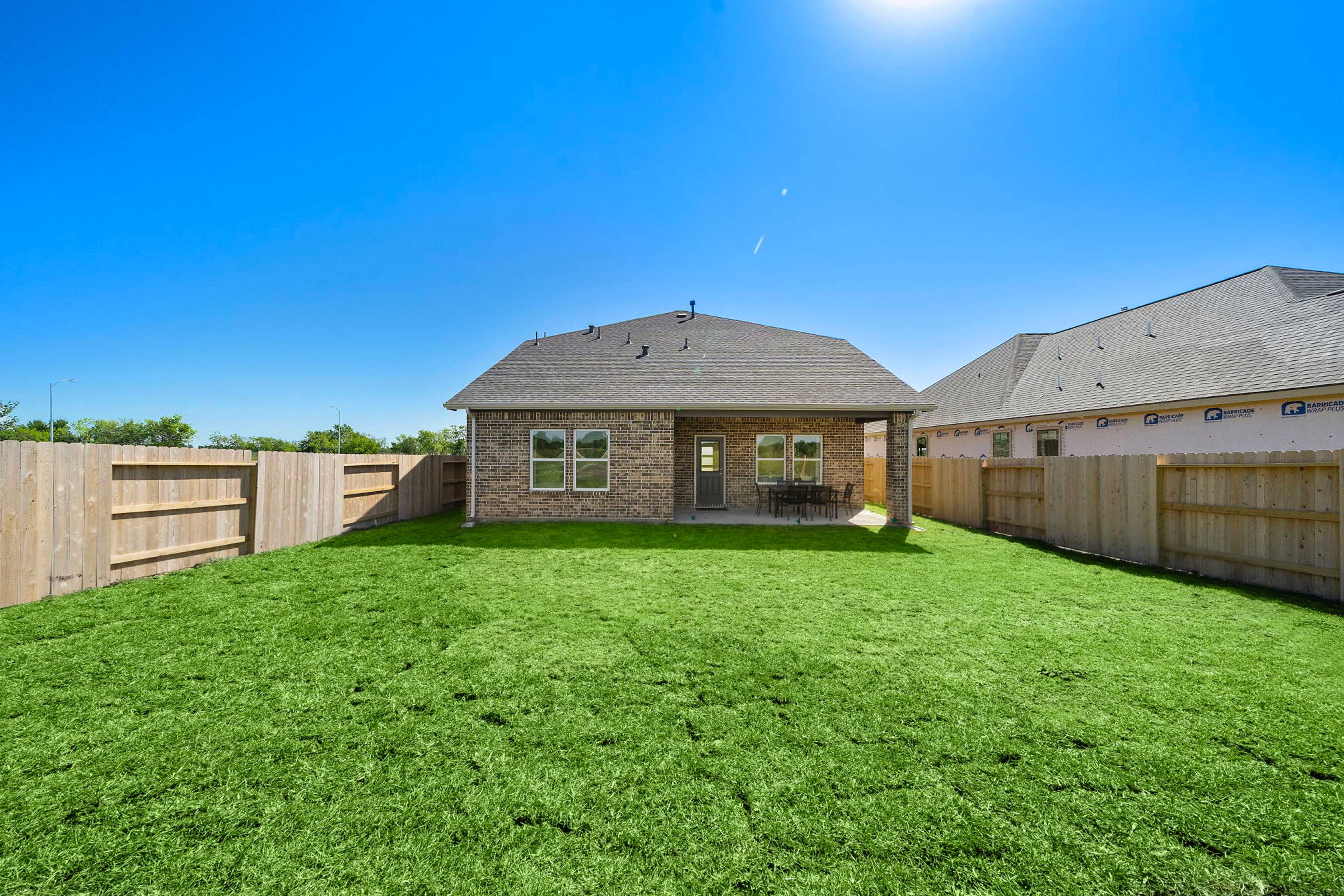 Fenced backyard with lush green lawn, covered patio seating, and single-story home at Emberly in Beasley, Texas