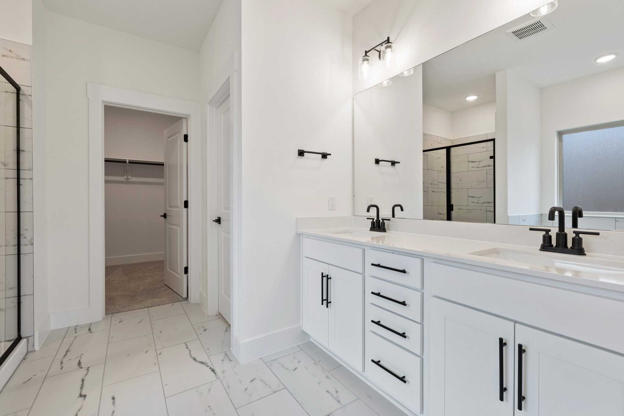 Modern master bathroom featuring double vanity with black faucets, frameless shower, and walk-in closet in Davidson Homes The Rockford C, Josephine, TX