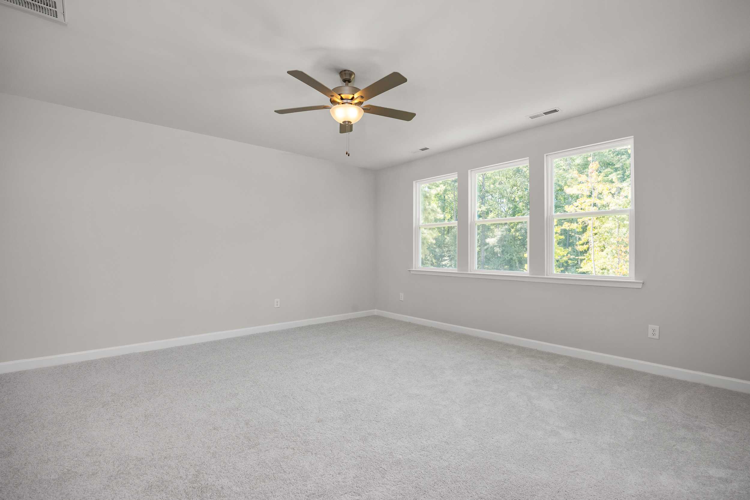 Spacious empty bedroom in The Aspen C home with light gray walls, neutral carpet, ceiling fan, and large triple windows with tree views