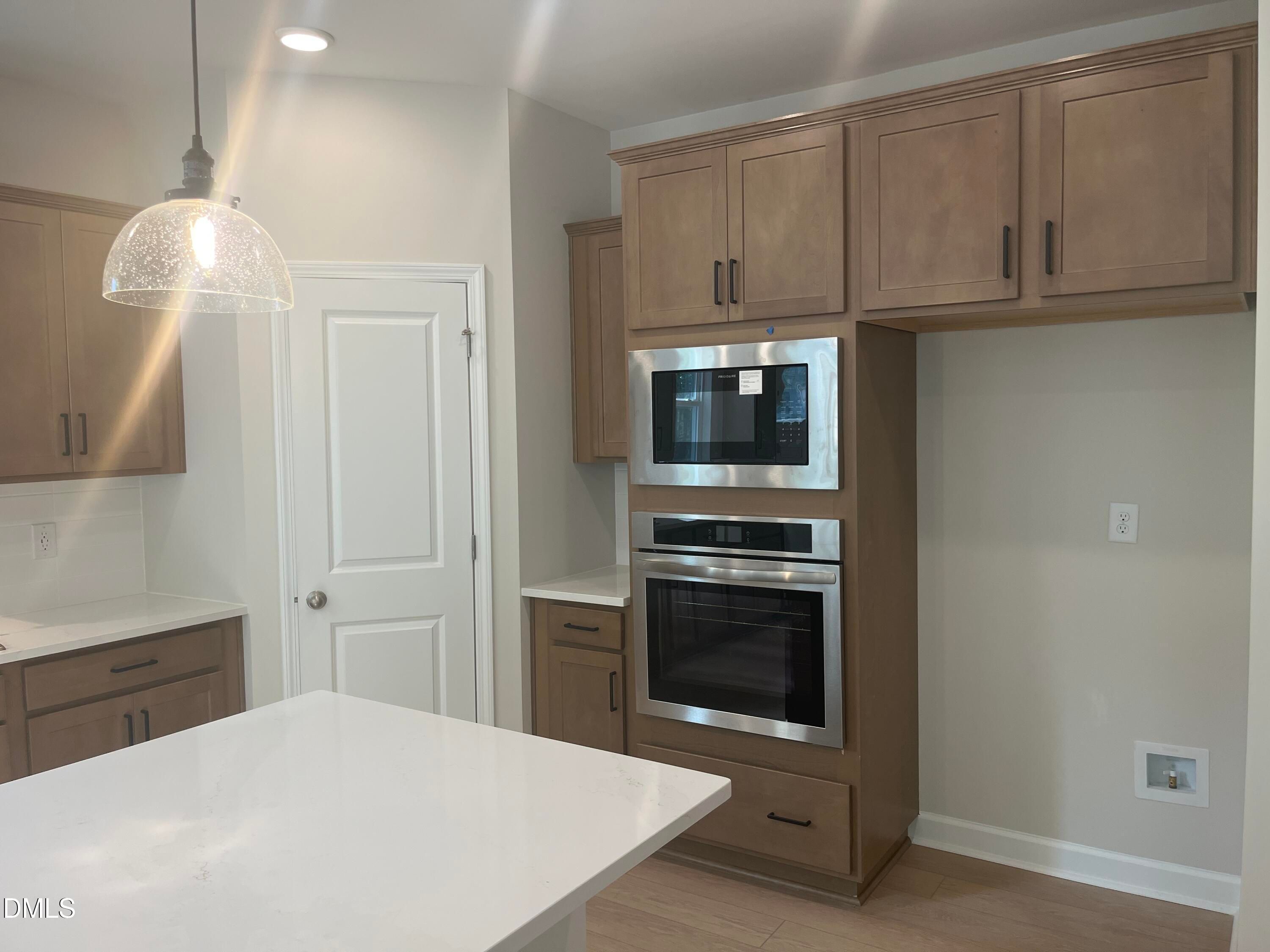 Modern kitchen with white quartz island, brown cabinets, stainless double wall oven, pendant light in The Ashport L, Wake Forest, NC
