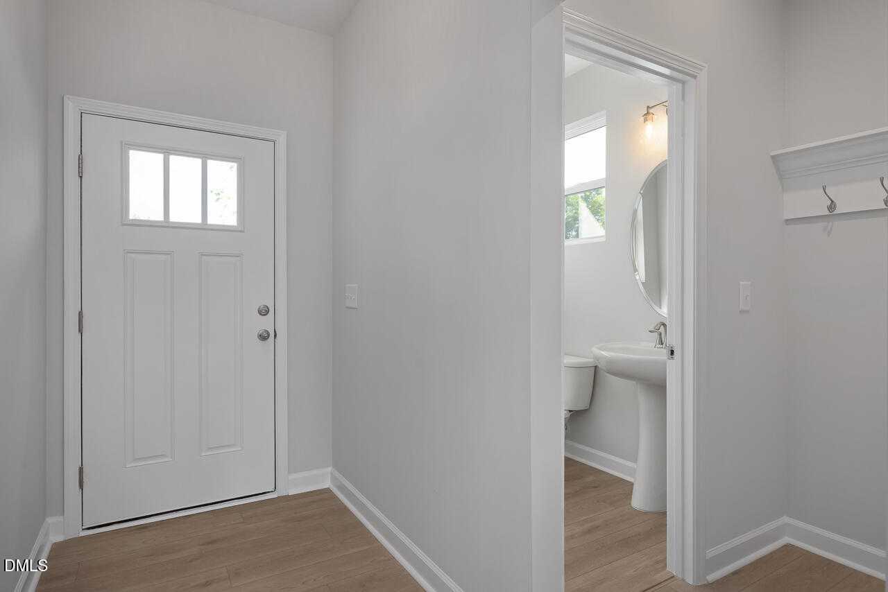 Bright entryway with glass-paneled white door, coat hooks, and adjacent powder room with pedestal sink in Davidson Homes The Graham, Fuquay-Varina, NC