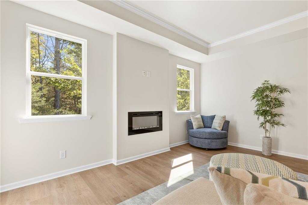 Cozy living room with linear gas fireplace, sunlit windows overlooking trees, blue tufted armchair, and hardwood floors in Davidson Homes The Daphne B, Loganville GA