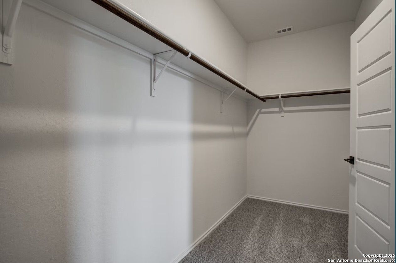 Spacious walk-in closet with wooden hanging rods, shelves, white walls, and gray carpet in Davidson Homes The Daphne H, Seguin, Texas
