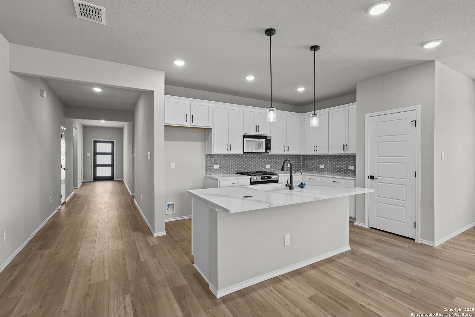 Open-concept kitchen with white cabinets, quartz island, stainless appliances, and adjacent hallway in The Daphne K, Comanche Ridge, San Antonio