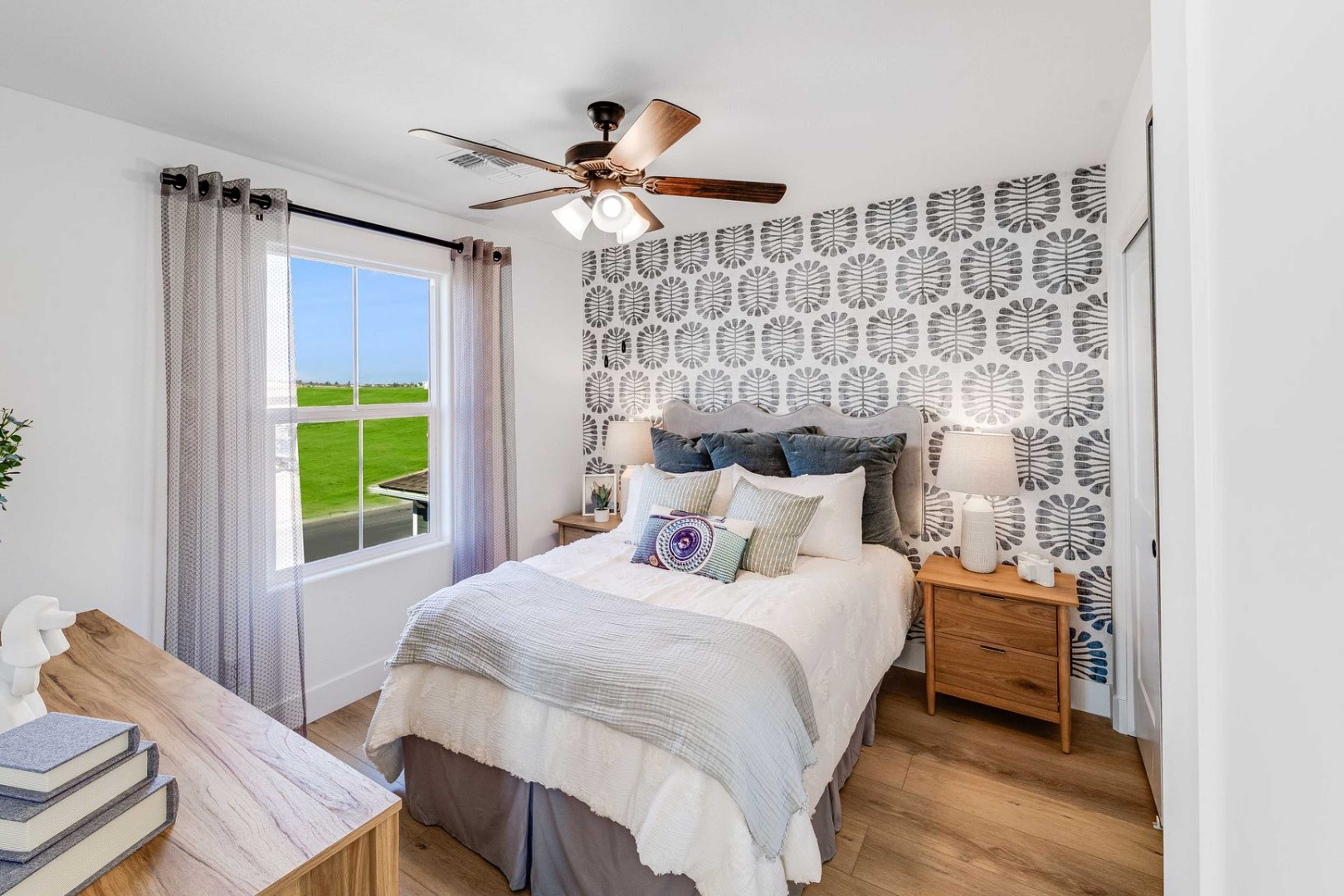Cozy secondary bedroom in The Wilmington home with gray patterned accent wall, queen bed in neutral linens, wooden dresser and nightstand, large window to green field view, ceiling fan