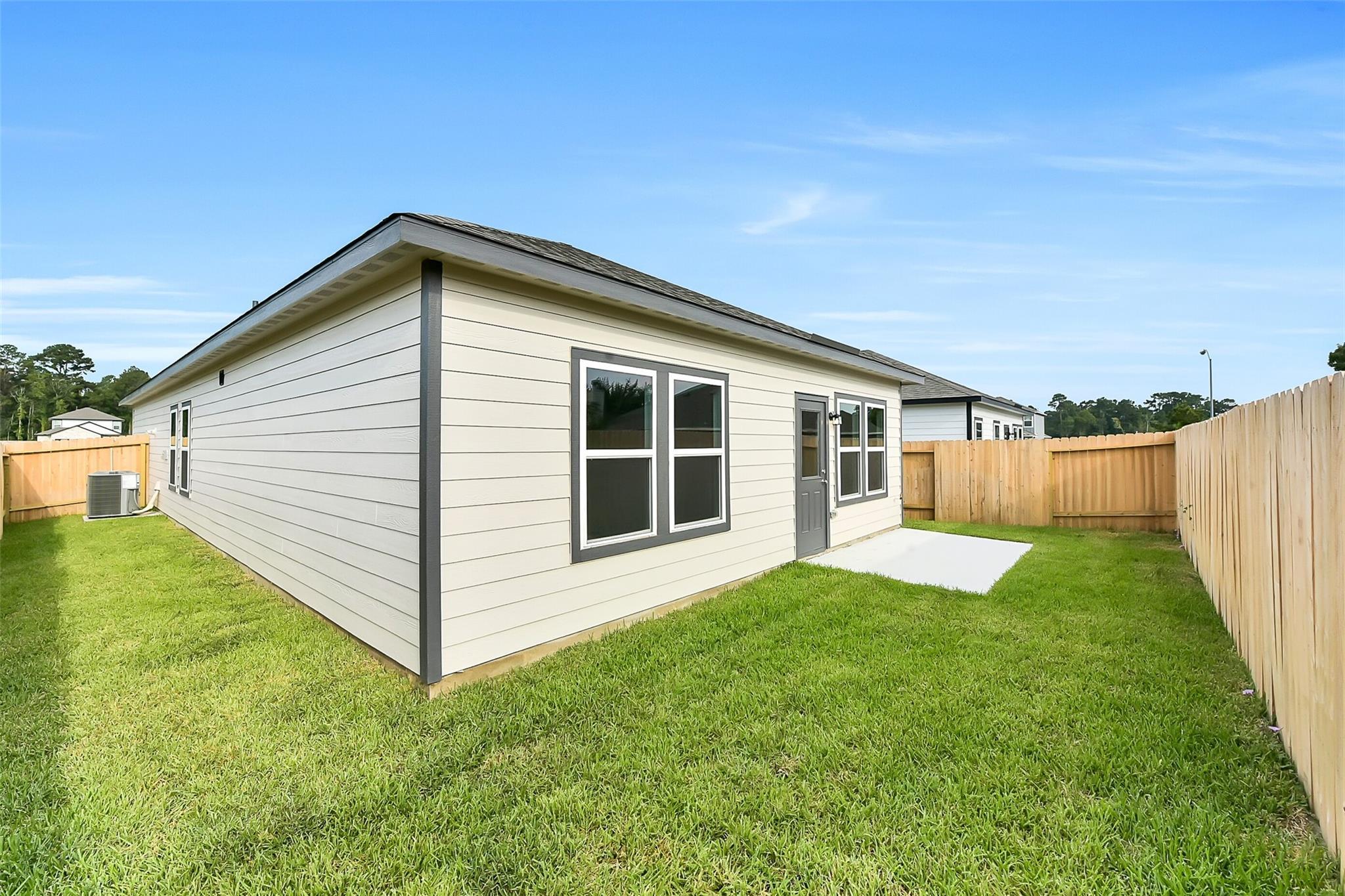 Modern beige single-story home exterior with side door, large windows, lush green yard, and wooden fence in The Villages at WestPointe, Dayton, Texas