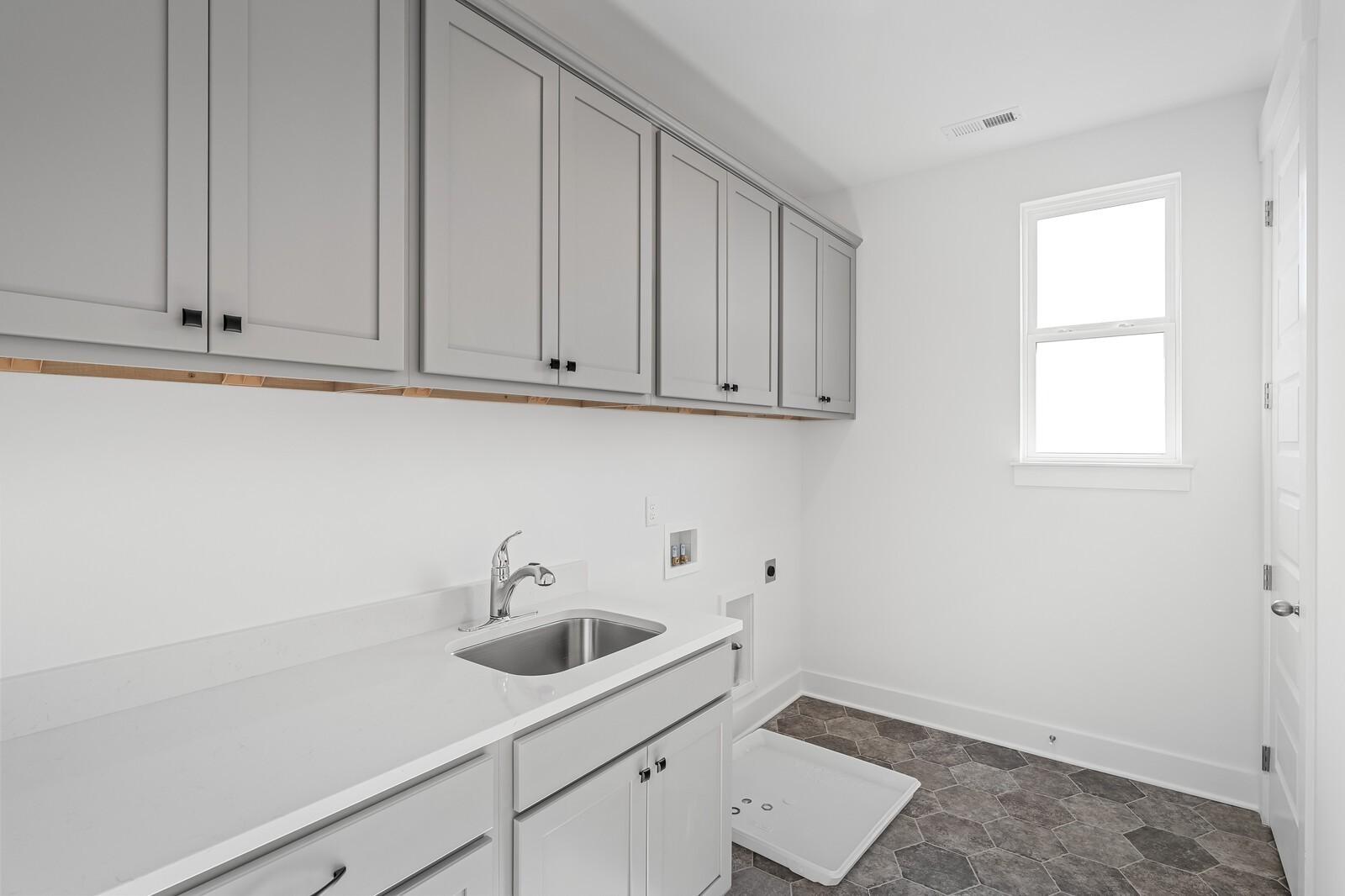 Modern laundry room with gray upper cabinets, white sink countertop, and washer-dryer area in Davidson Homes The Alston A, Murfreesboro, TN