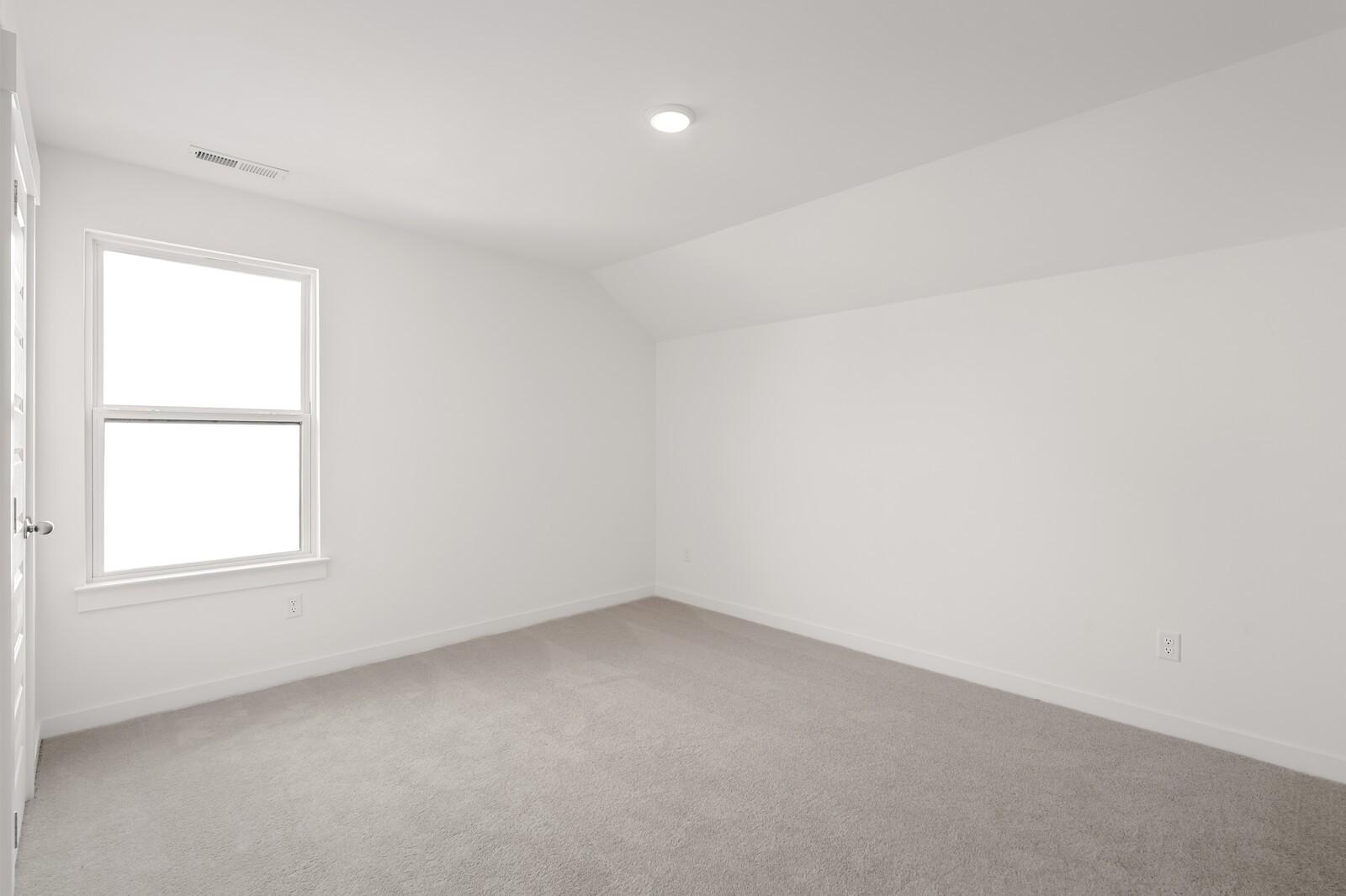 Bright empty bedroom with white walls, sloped ceiling, large window, and beige carpet in Davidson Homes The Ash C, Mt. Juliet