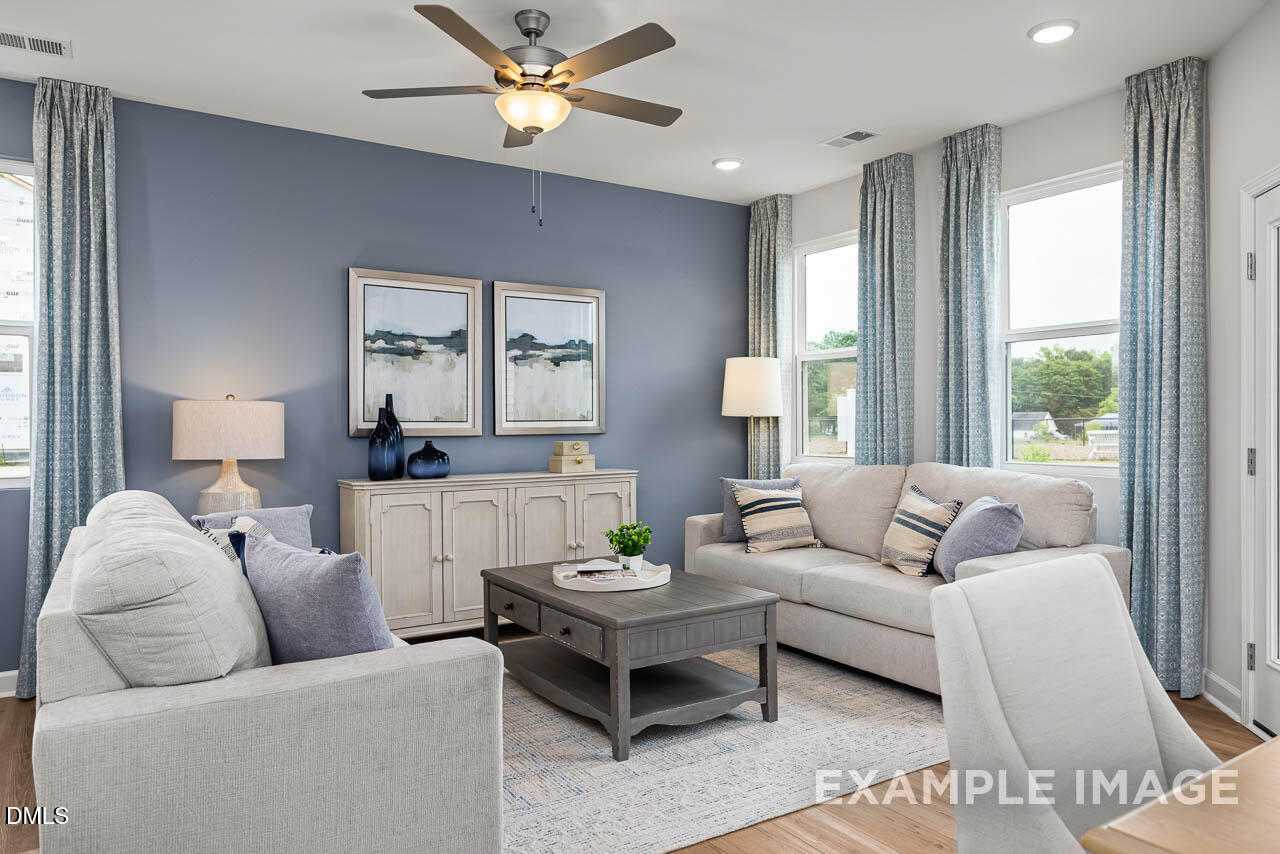 Cozy living room with blue walls, white sofas, ceiling fan, and large windows in Davidson Homes The Graham, Fuquay-Varina, NC