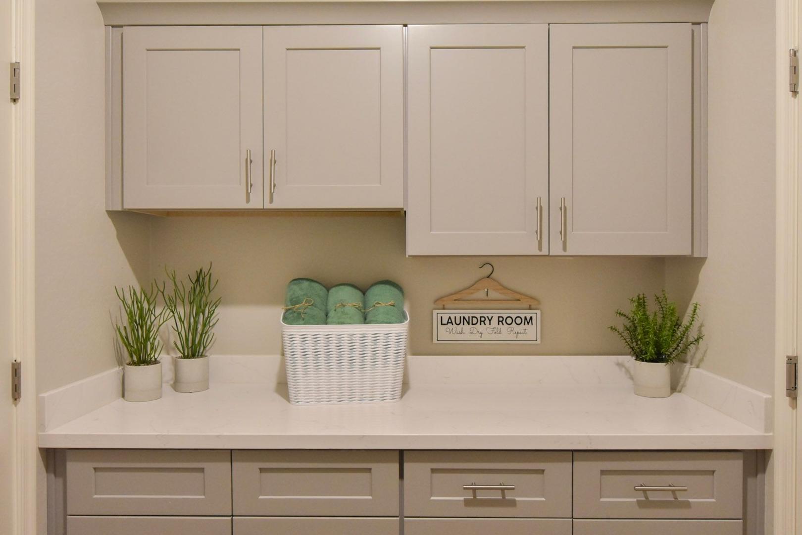 Spacious laundry room in The Harmony B featuring white shaker cabinets, quartz countertop, green towels in wicker basket, and potted plants