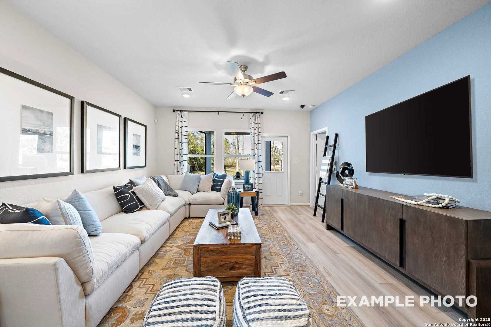 Cozy modern living room with L-shaped white sofa, wall-mounted TV, ceiling fans, and wooden coffee table in Davidson Homes The San Marcos D, San Antonio