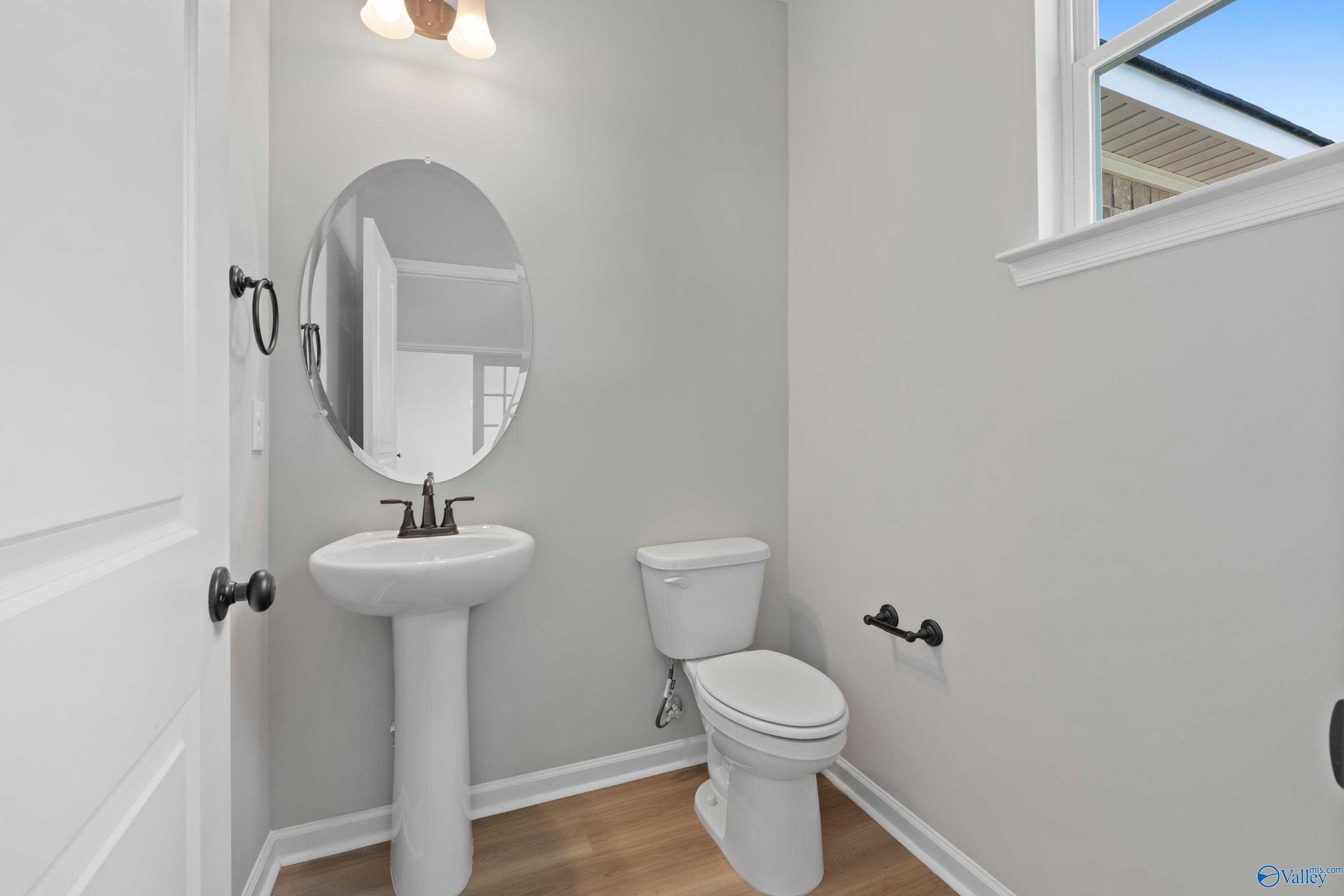 Elegant powder room featuring pedestal sink, oval mirror, toilet, gray walls, and hardwood floors in Davidson Homes The Dorado, Huntsville, Alabama