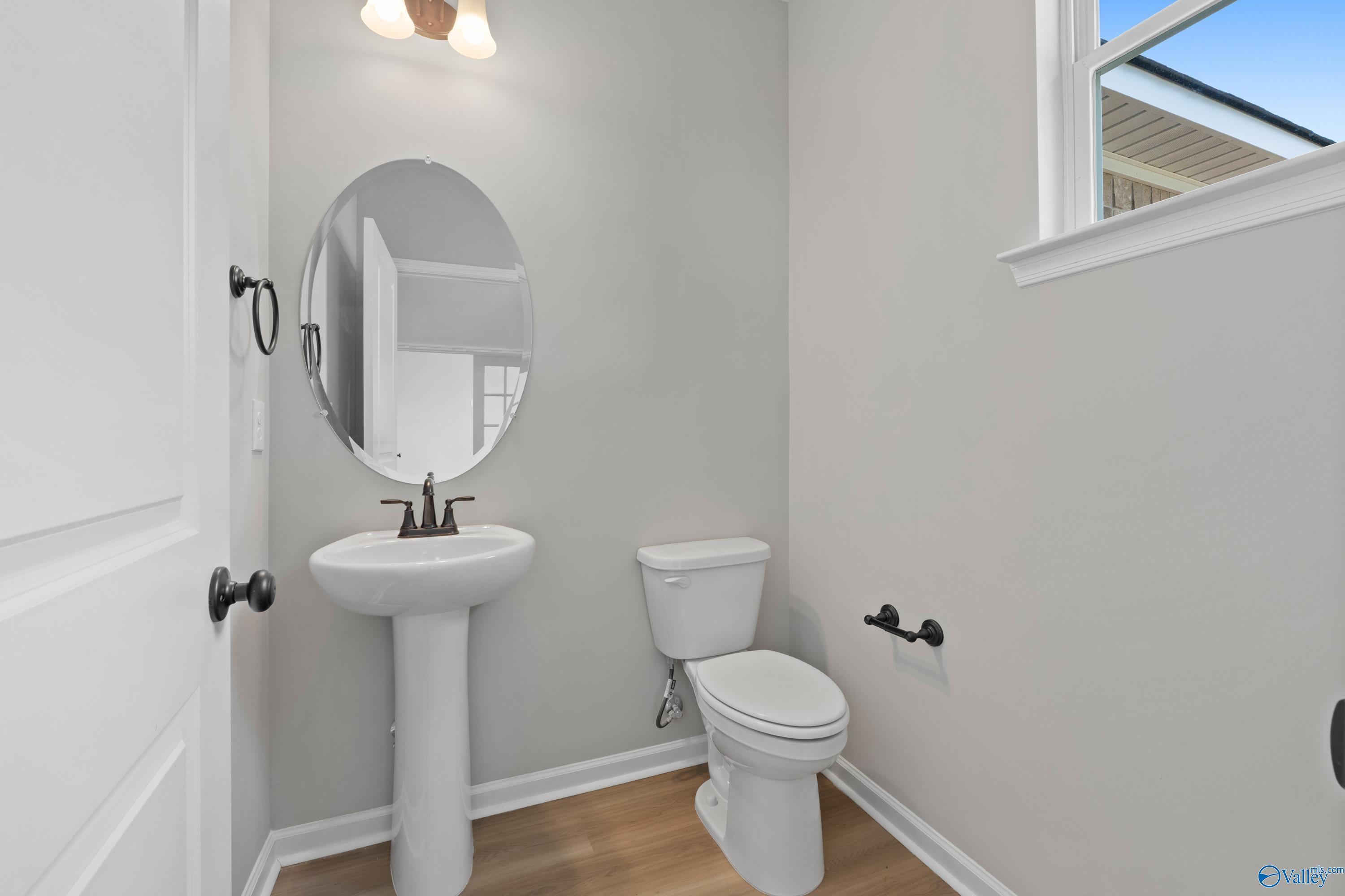 Elegant powder room featuring pedestal sink, oval mirror, toilet, gray walls, and hardwood floors in Davidson Homes The Dorado, Huntsville, Alabama