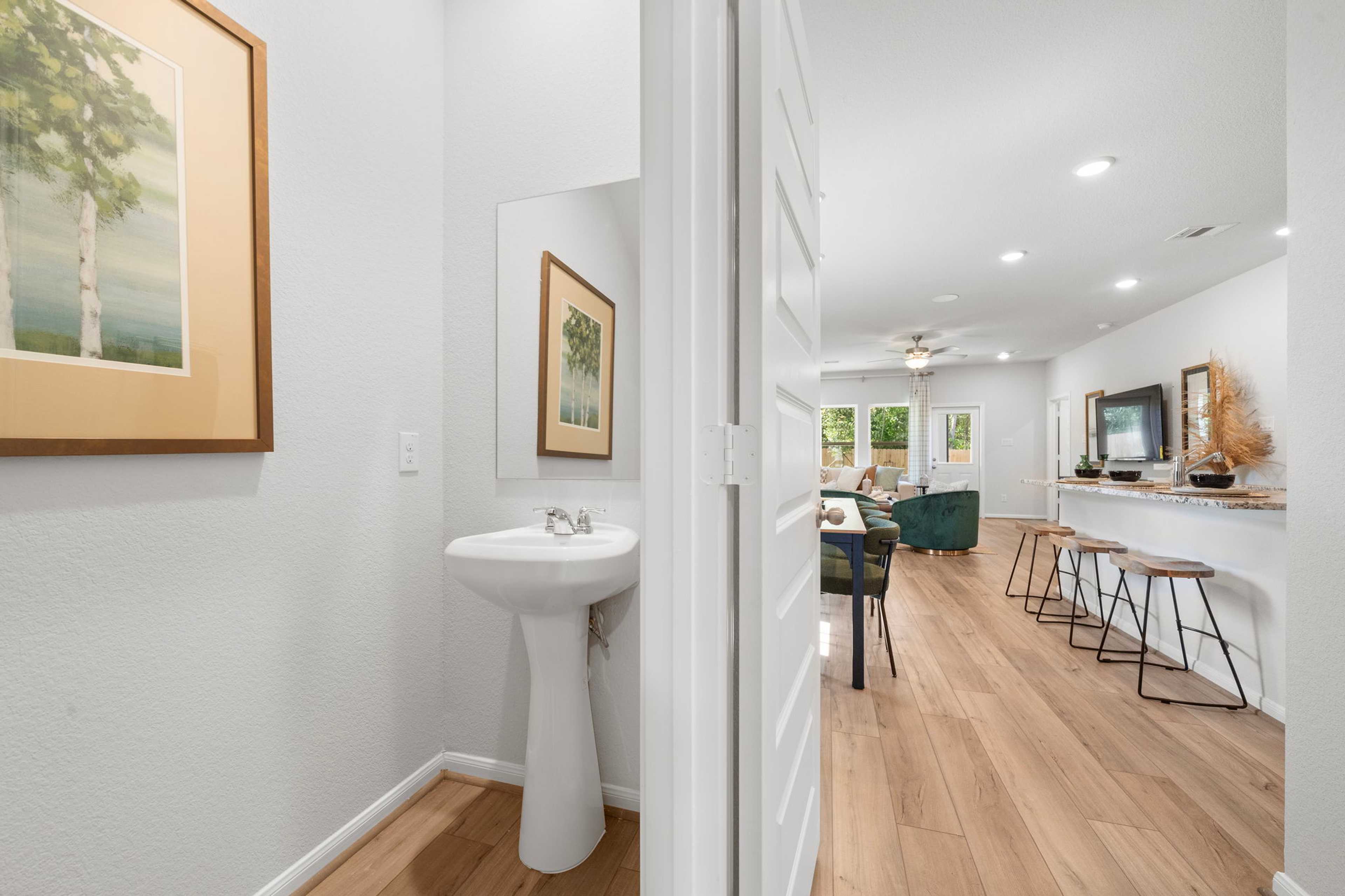 Open-concept powder room with pedestal sink and birch tree artwork opening to kitchen bar on hardwood floors in Caney Creek Place, Conroe TX by Davidson Homes