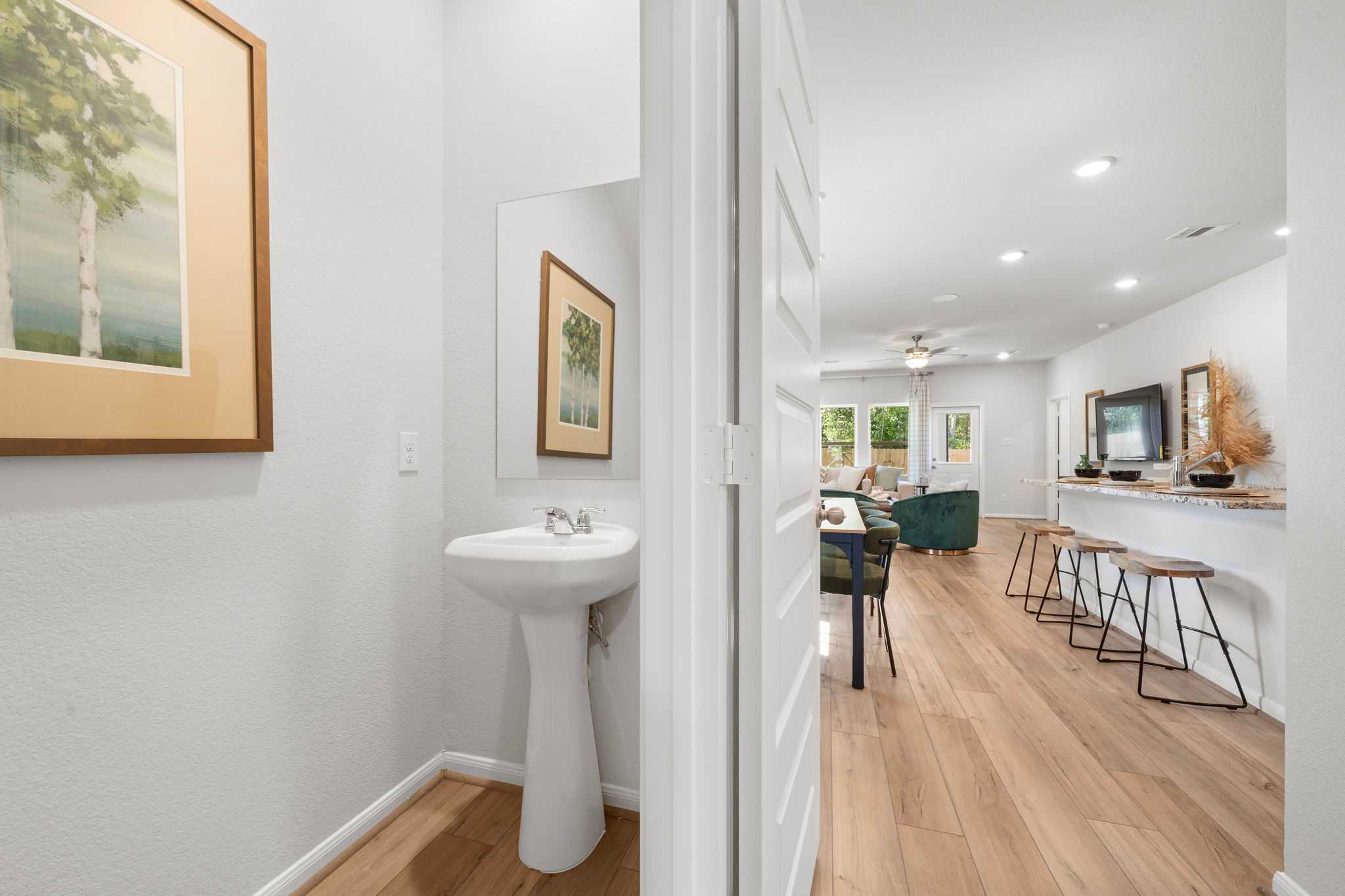 Open-concept powder room with pedestal sink and birch tree artwork opening to kitchen bar on hardwood floors in Caney Creek Place, Conroe TX by Davidson Homes