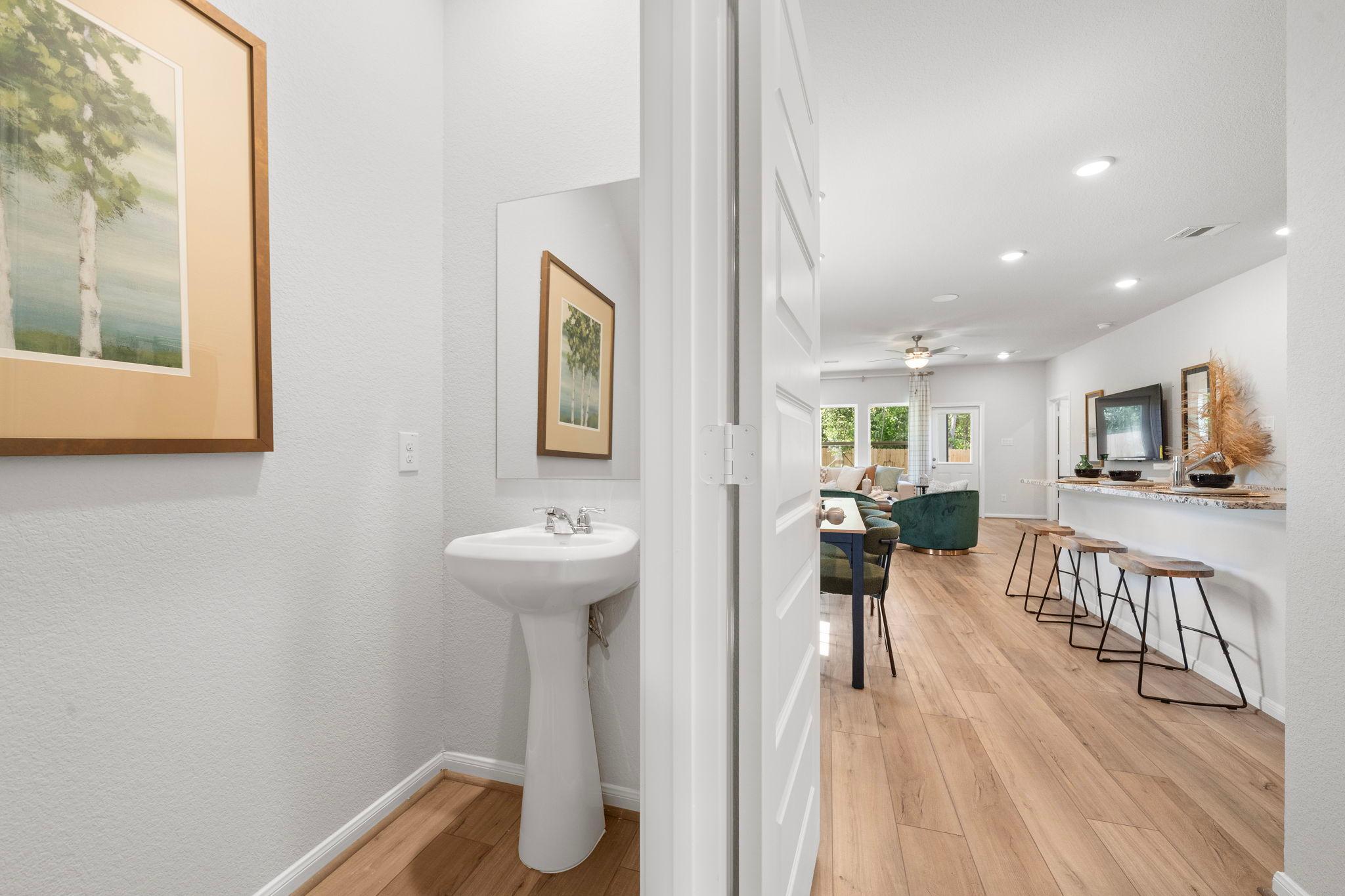 Open-concept powder room with pedestal sink and birch tree artwork opening to kitchen bar on hardwood floors in Caney Creek Place, Conroe TX by Davidson Homes