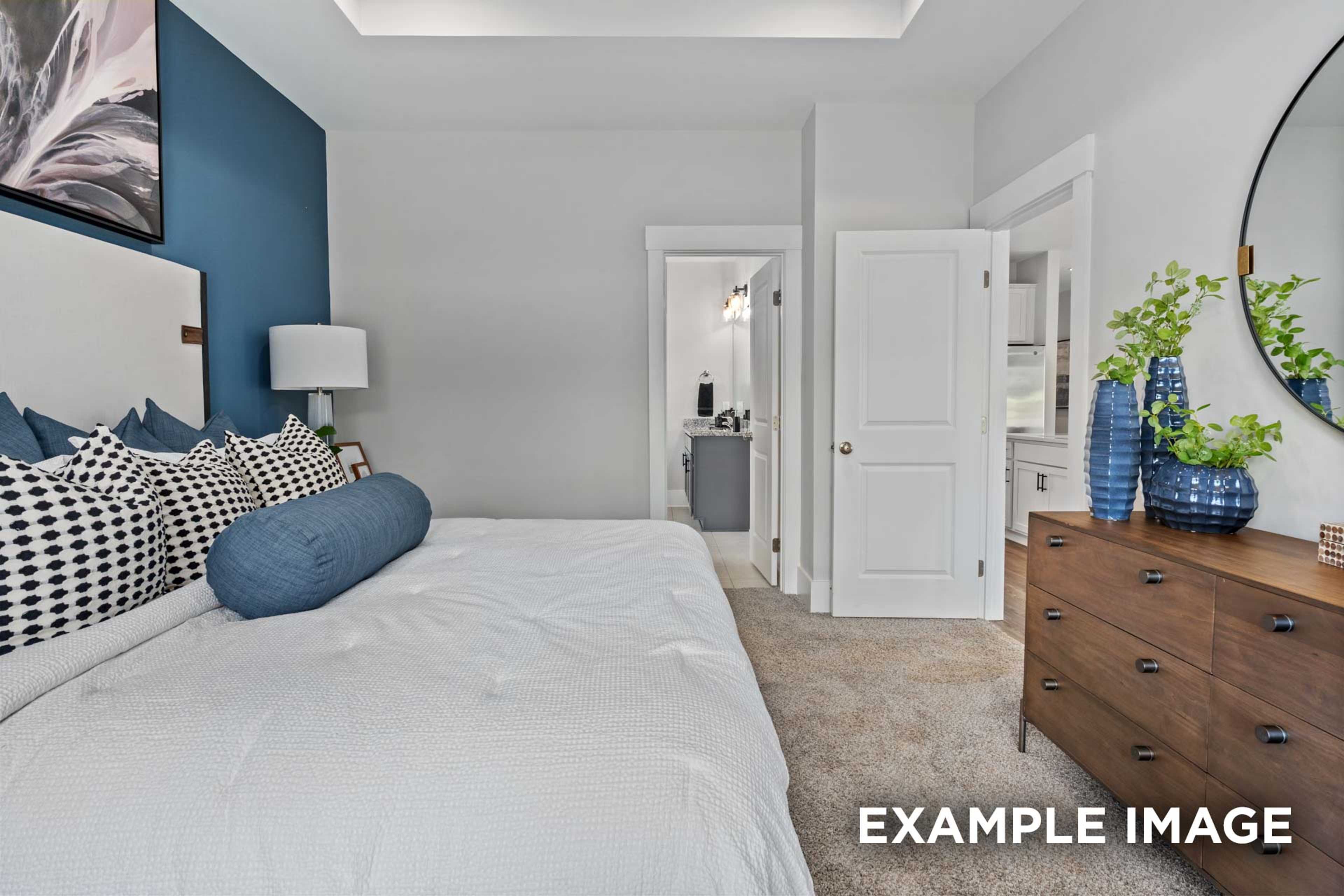 Spacious master bedroom in The Franklin C with white duvet bed, blue pillows, wooden dresser, round mirror, and en-suite bath access