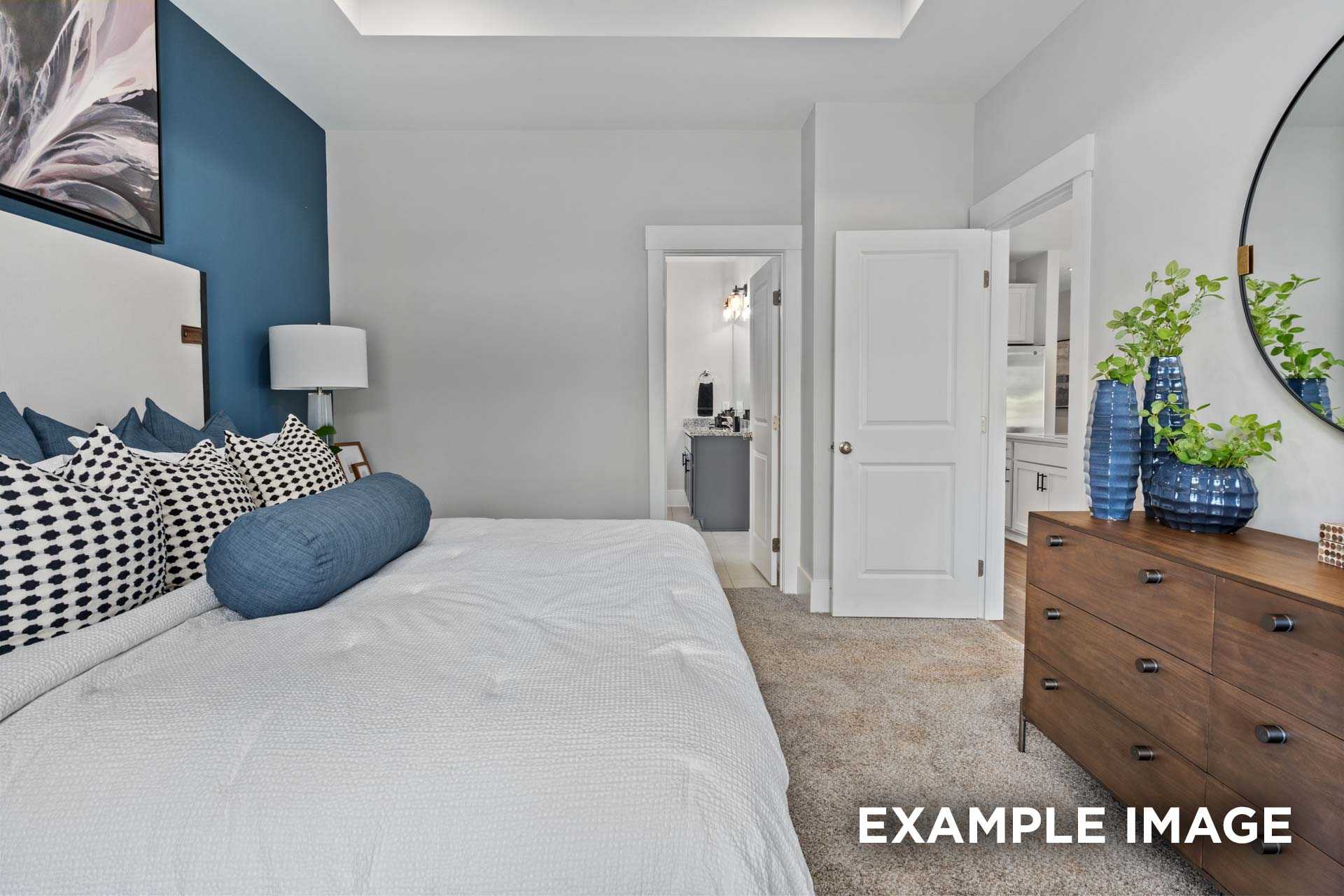 Spacious master bedroom in The Franklin C with white duvet bed, blue pillows, wooden dresser, round mirror, and en-suite bath access