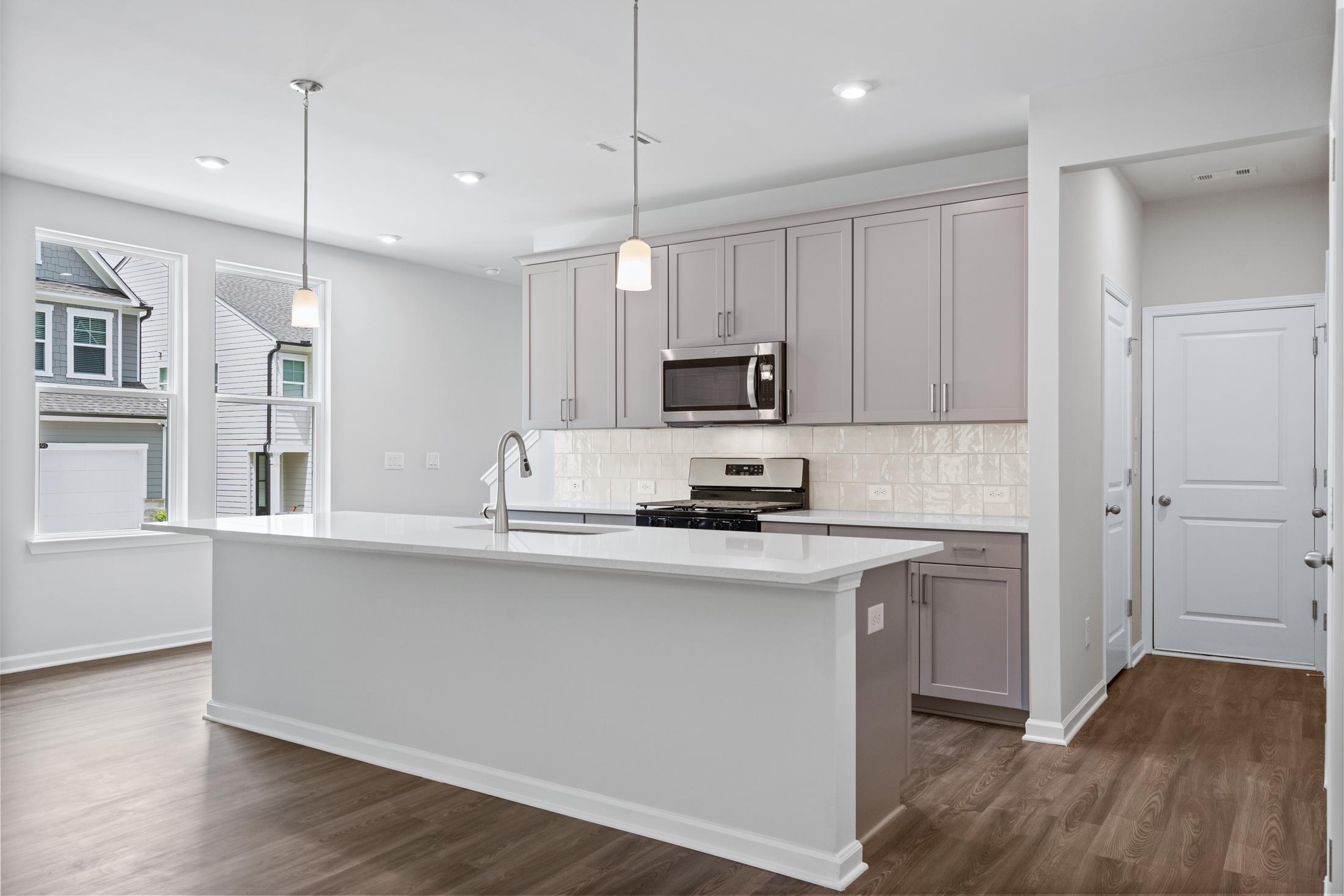 Bright kitchen in The Cary A with large white island, light gray cabinets, stainless steel appliances, subway tile backsplash, and hardwood floors