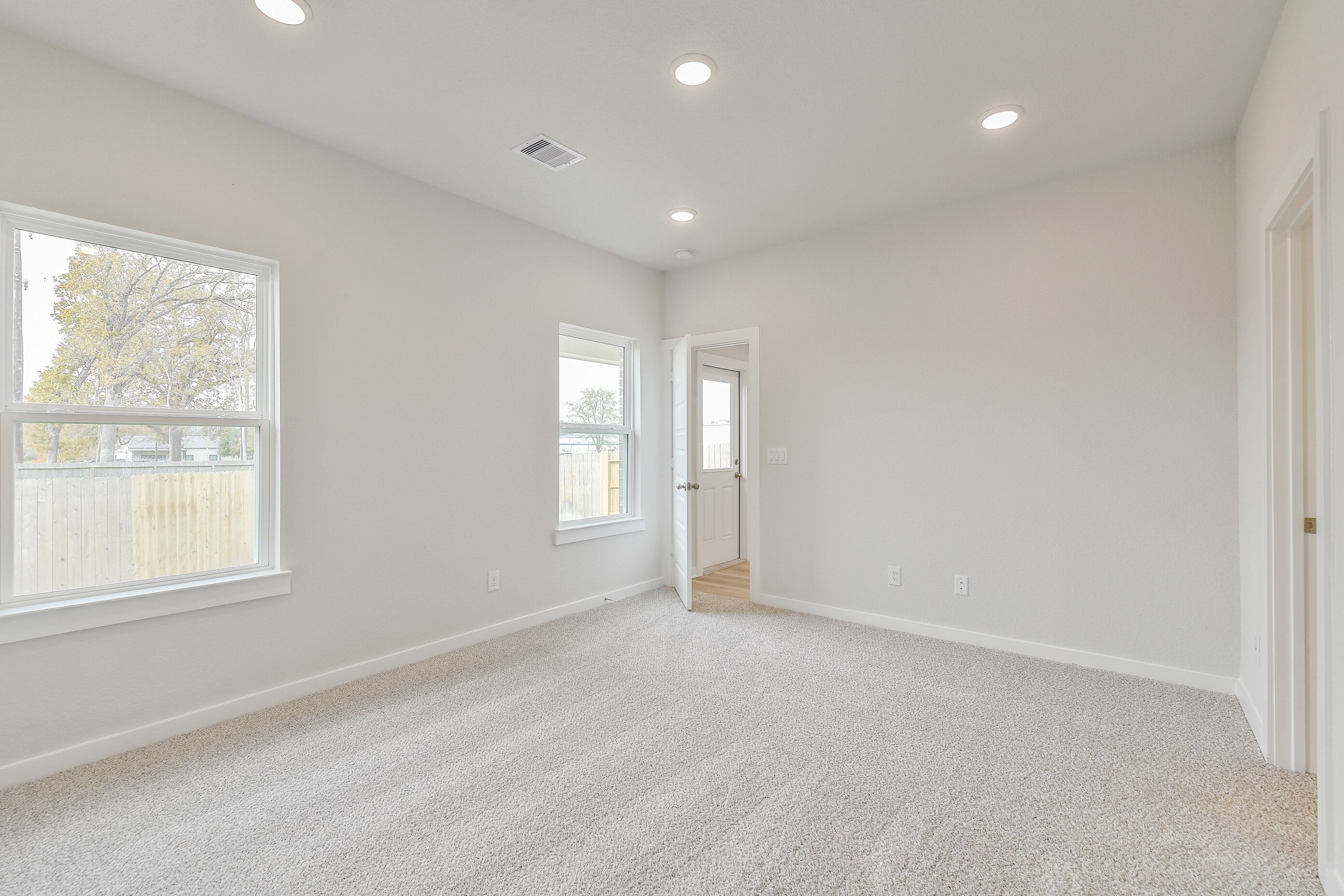 Bright empty bedroom with neutral gray walls, beige carpet, recessed lights, and large windows overlooking trees in Davidson Homes The Colorado G, Magnolia Texas