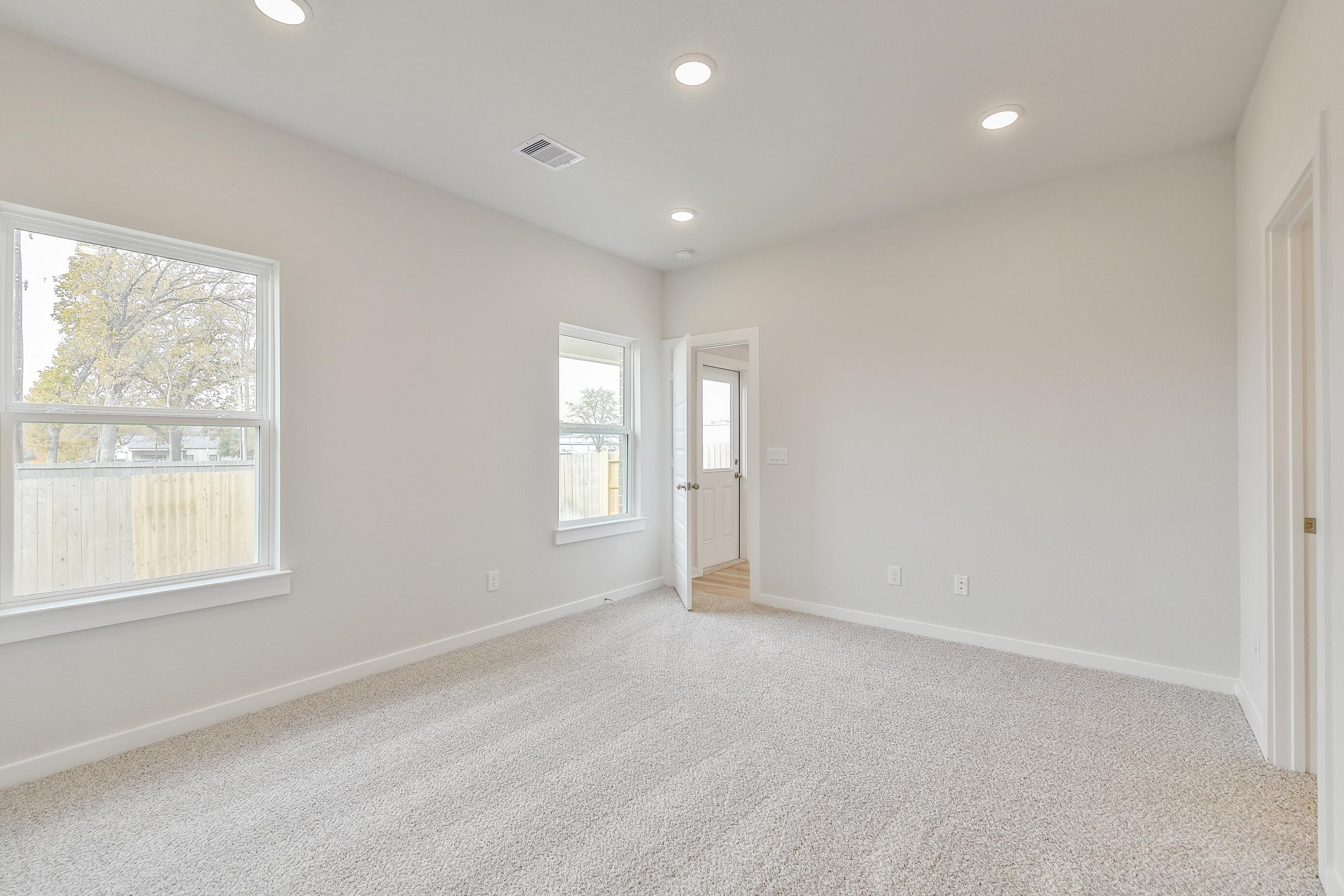 Bright empty bedroom with neutral gray walls, beige carpet, recessed lights, and large windows overlooking trees in Davidson Homes The Colorado G, Magnolia Texas
