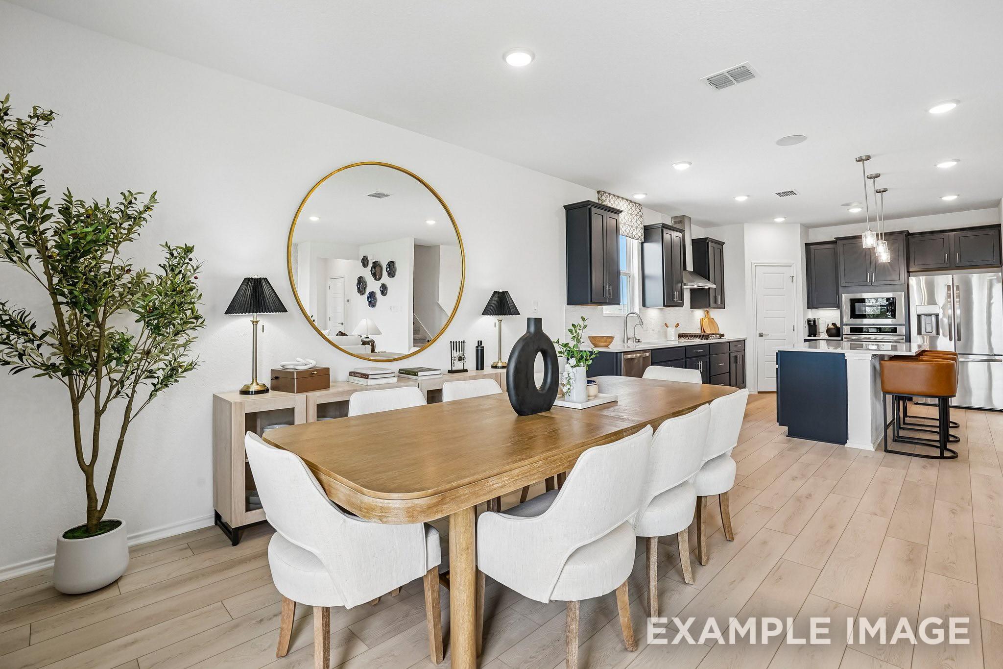 Open-concept dining area with wood table, white chairs, gold mirror, and dark cabinetry kitchen in The Ashport J, Crosby, Texas