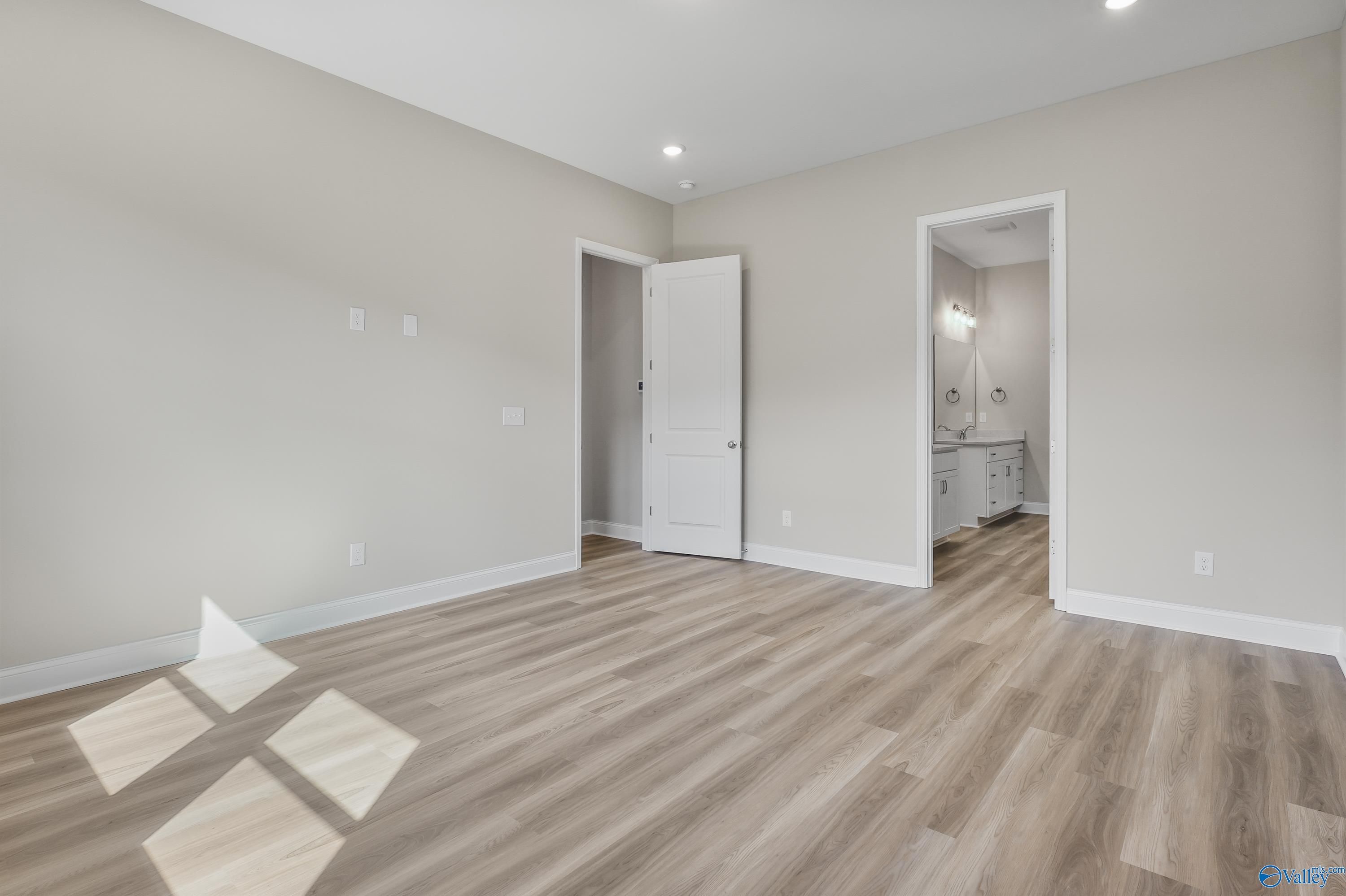 Bright bedroom with beige walls, light hardwood floors, and adjacent bathroom featuring white vanity in Evermore Homes The Haven, Owens Cross Roads, AL