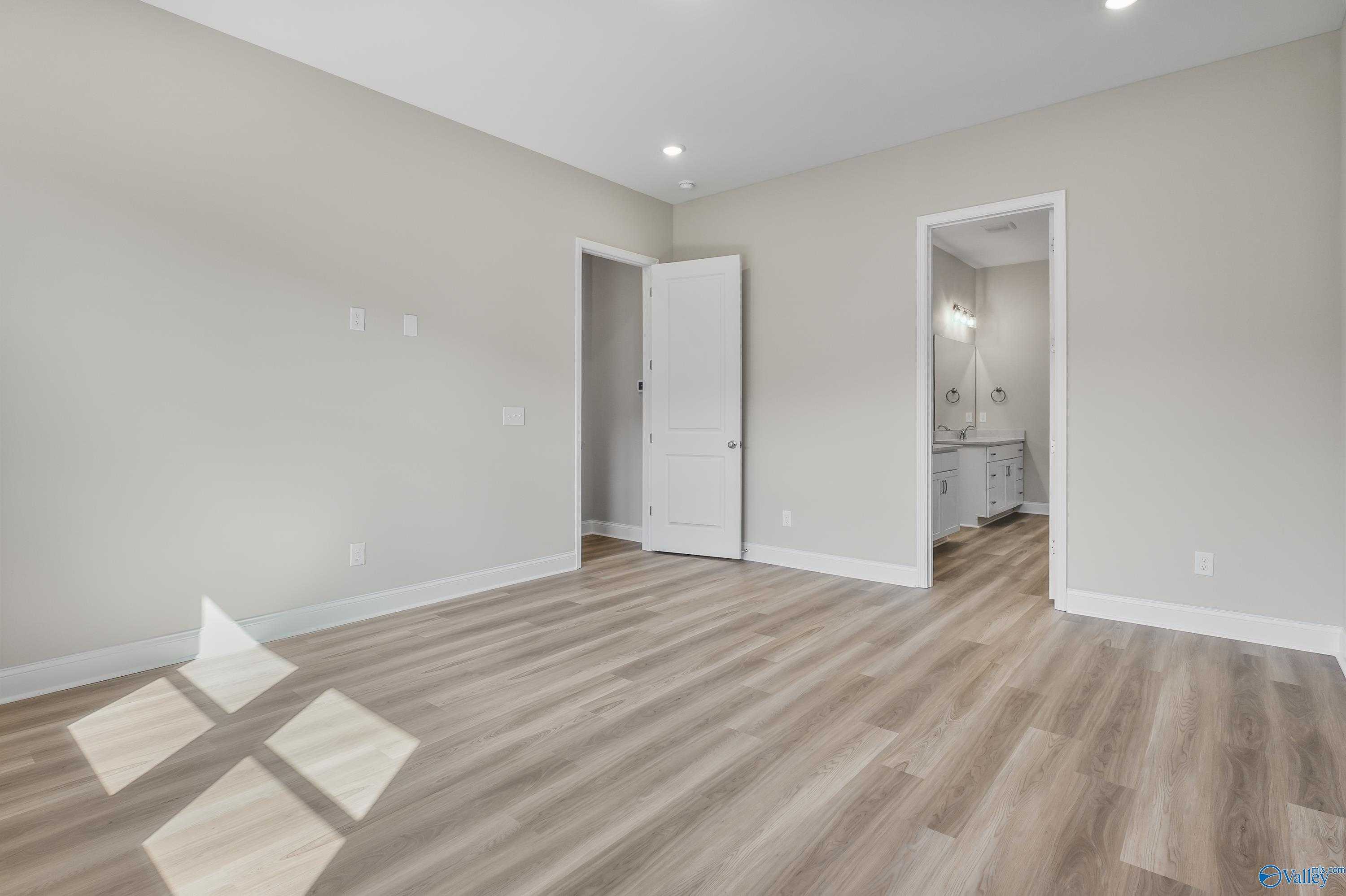 Bright bedroom with beige walls, light hardwood floors, and adjacent bathroom featuring white vanity in Evermore Homes The Haven, Owens Cross Roads, AL