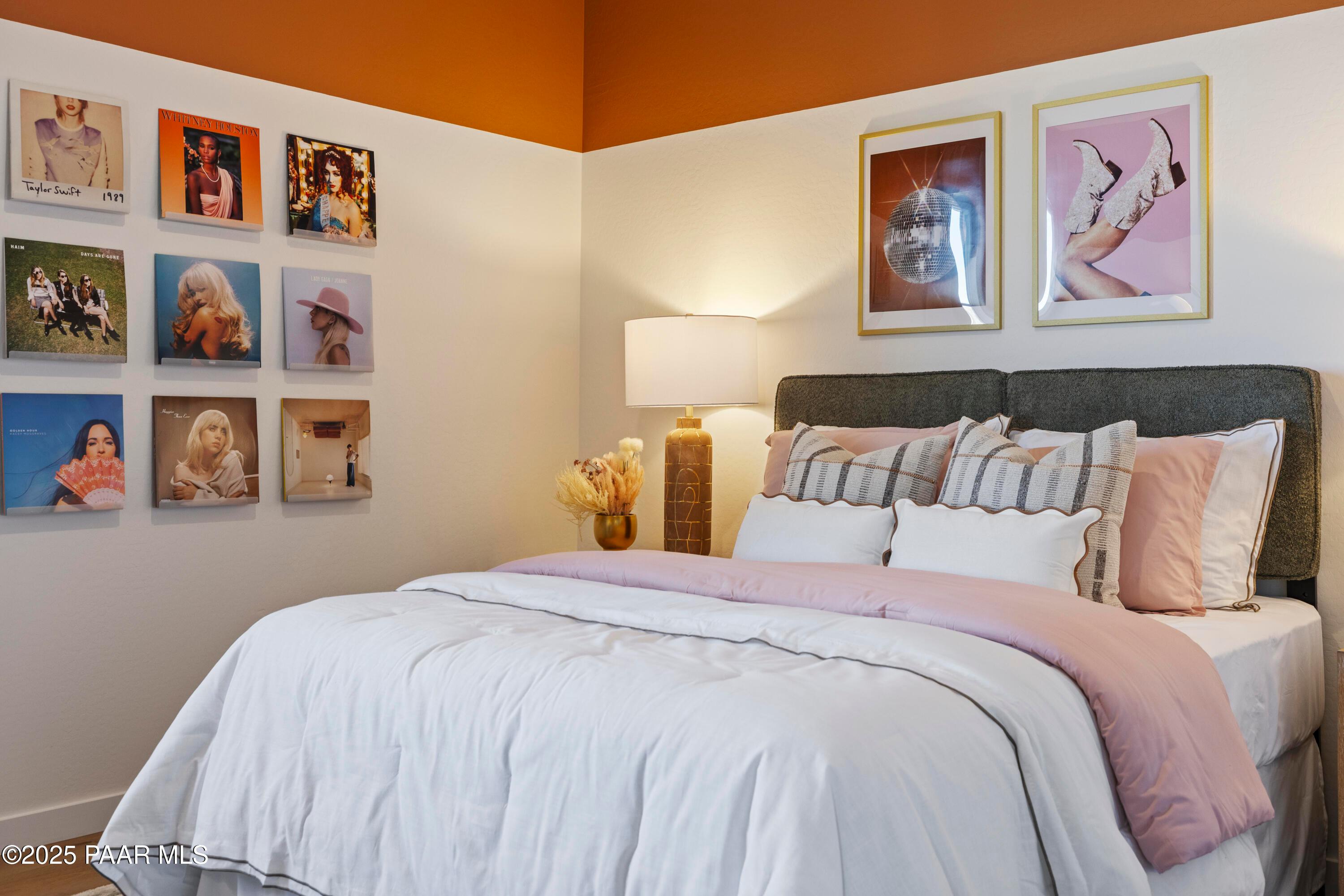Cozy bedroom with orange accent wall, framed photo gallery, tufted gray headboard, and plush white bedding in Davidson Homes The Blaze D, Prescott