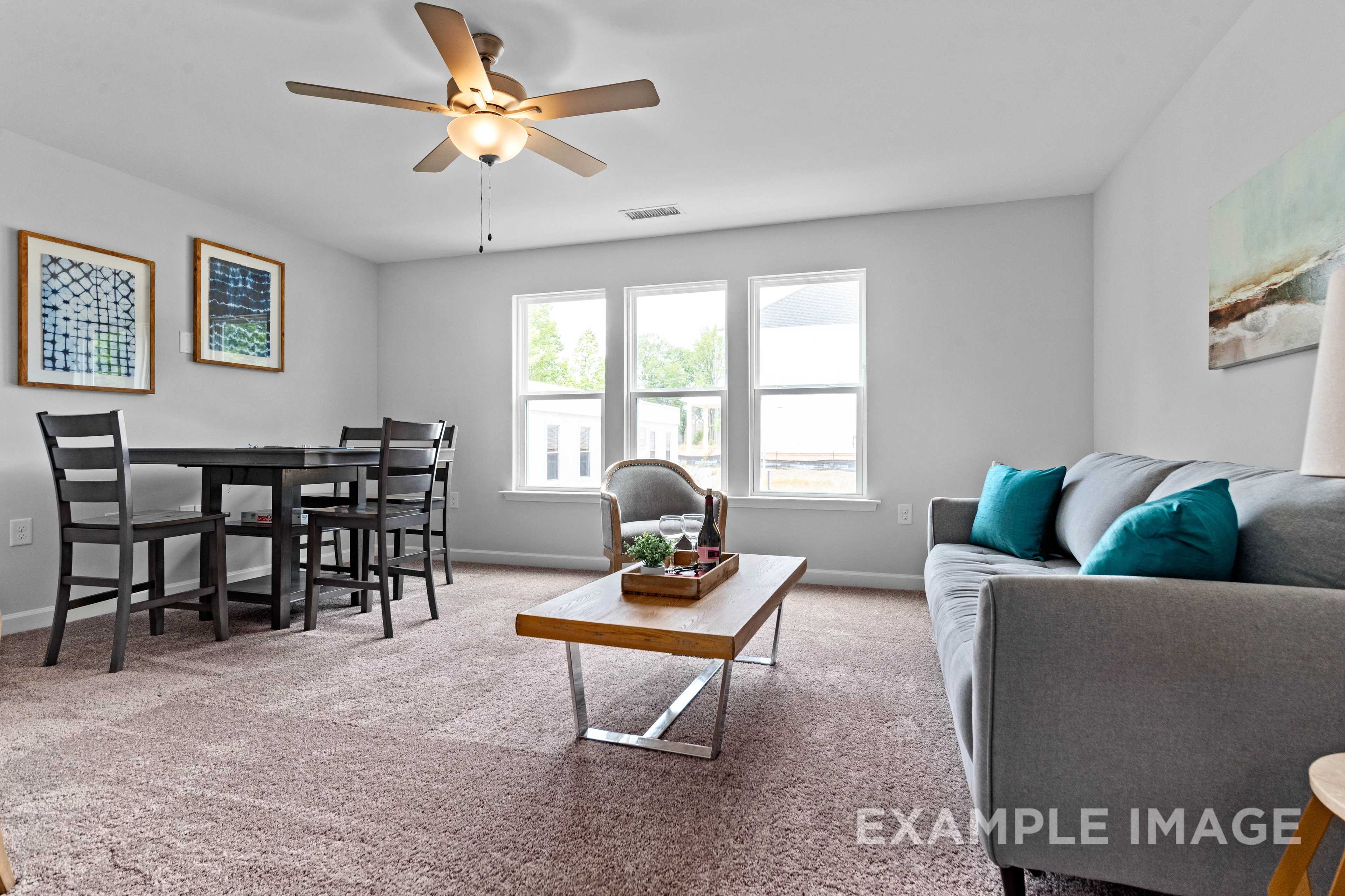 Open-concept living and dining area in The Ash C home, with modern wooden table, gray sofa, teal pillows, and large windows