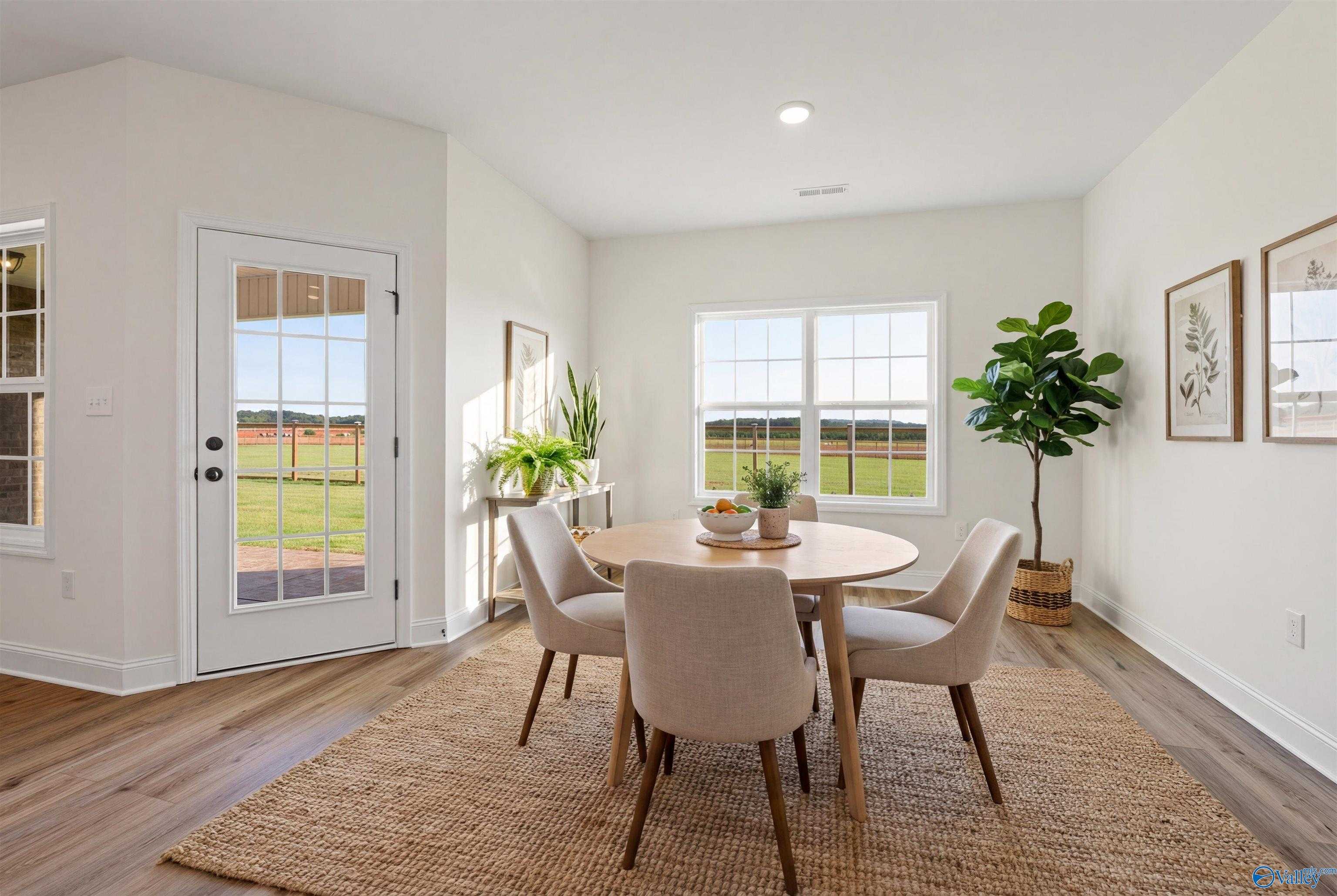 Bright dining room with round wood table, beige chairs, fiddle leaf fig, and large windows overlooking fields in The Rockford B, Toney, Alabama