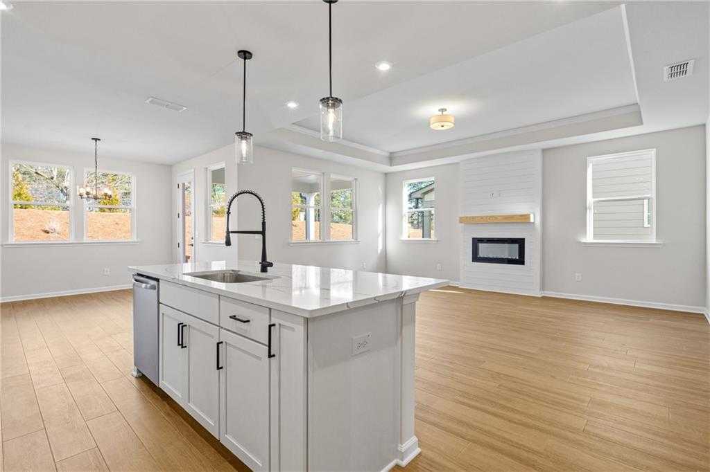 Modern open-concept kitchen with white island, stainless dishwasher, and pendant lights opening to family room with gas fireplace in The Edison A, Loganville, GA