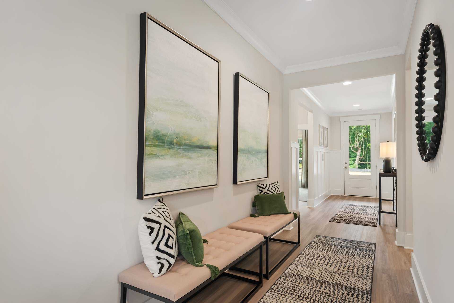 Elegant hallway in Durham Farms home, Harvest Alabama featuring abstract green wall art, tufted bench with pillows, and hardwood floors