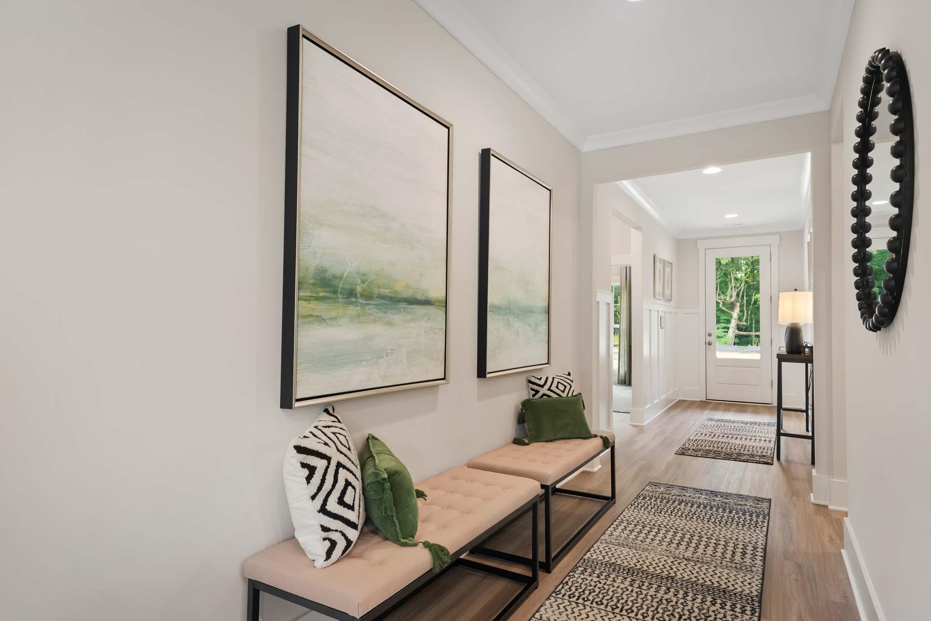 Elegant hallway in Durham Farms home, Harvest Alabama featuring abstract green wall art, tufted bench with pillows, and hardwood floors